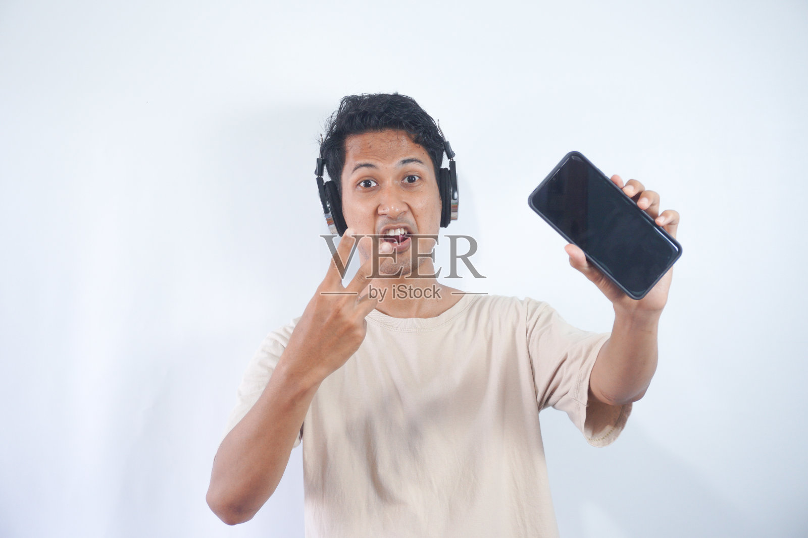 乐观的男人拿着模拟屏幕的智能手机，戴着耳机听着喜欢的音乐，穿着休闲的奶油色t恤，站在室内的半长镜头。人与技术的理念。照片摄影图片