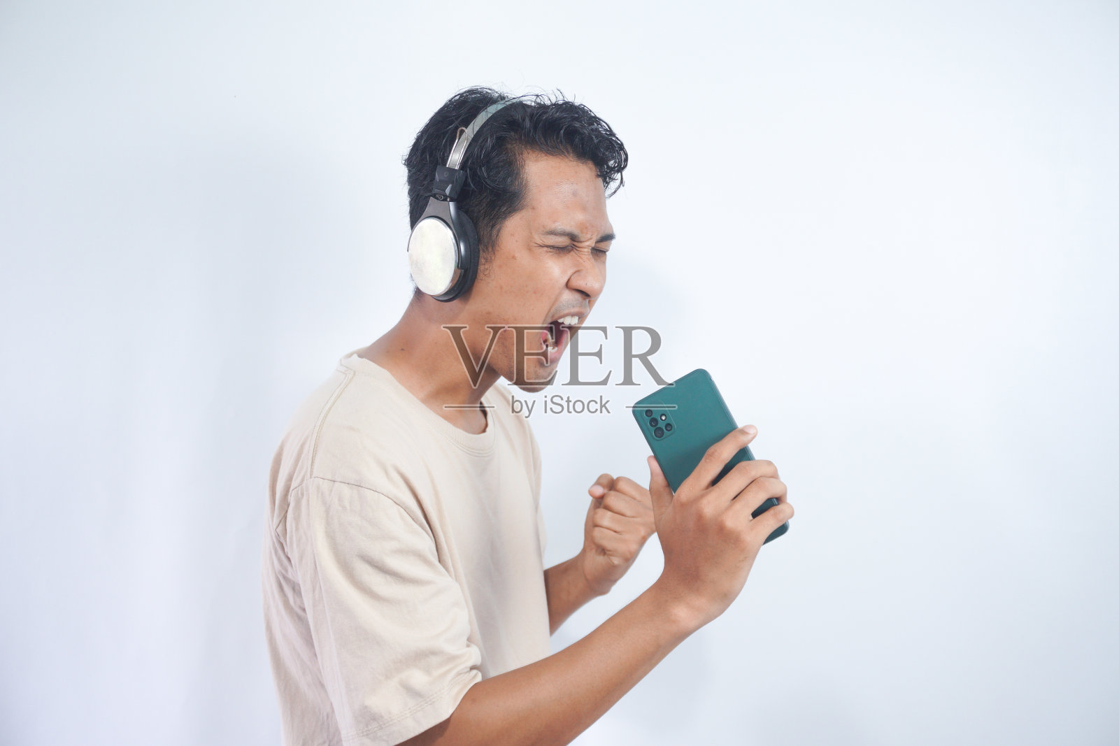 精力充沛的男人伴着音乐唱歌，动作积极，戴着耳机，穿着休闲的t恤，在白色的背景下摆姿势，张大嘴巴，享受生活，孤立在白色的工作室墙上照片摄影图片