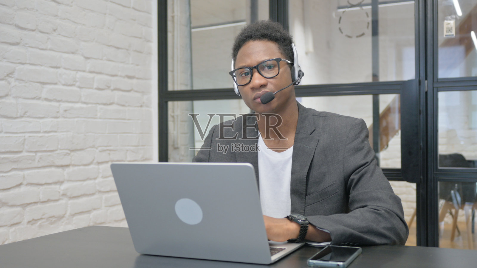 戴着耳机的非洲年轻人一边用笔记本电脑工作，一边看着摄像头照片摄影图片