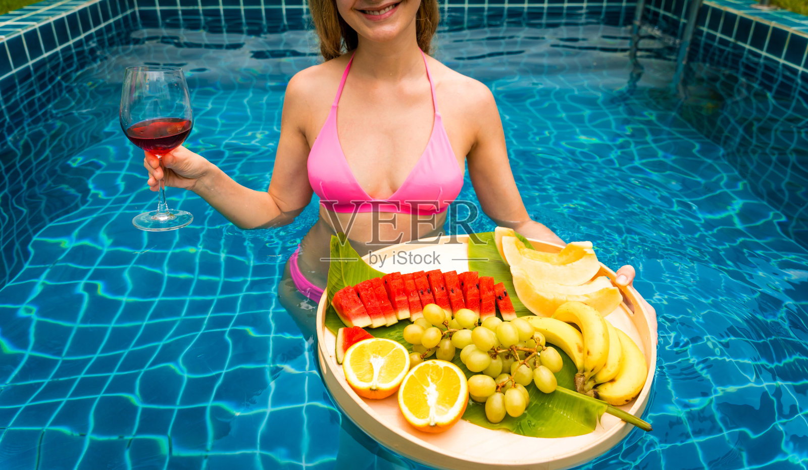 泳池里的美女端着红酒和漂浮的水果盘照片摄影图片