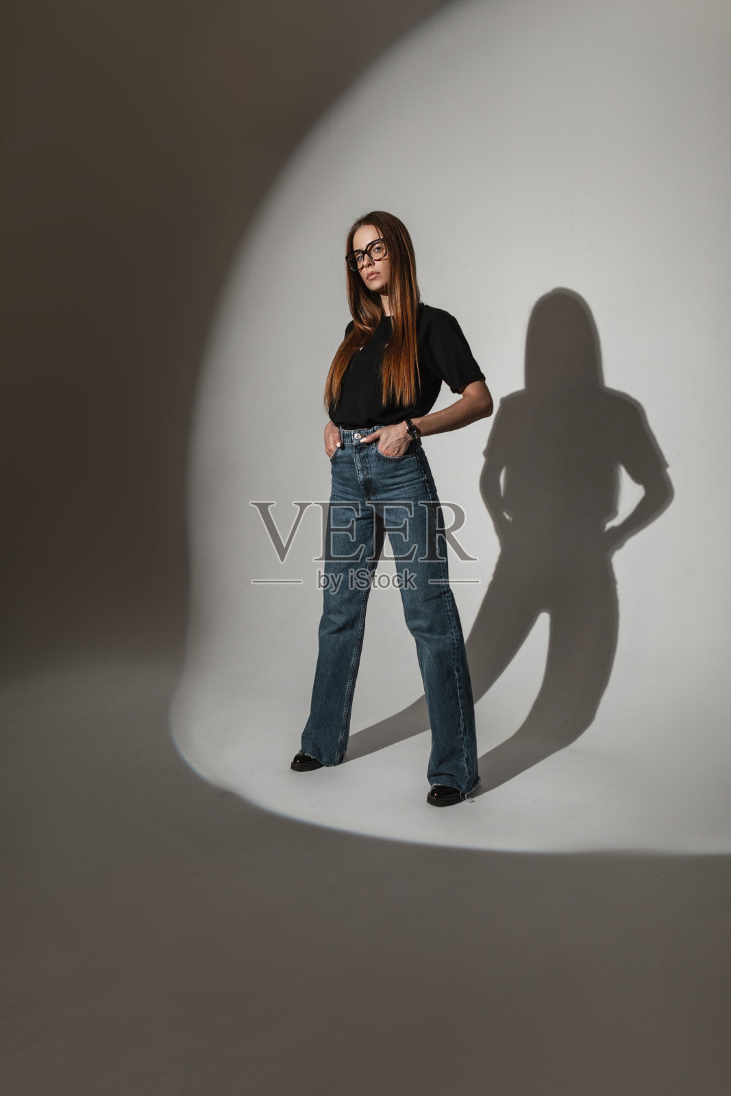 时尚的年轻女模特戴着眼镜，穿着时髦的衣服，黑色t恤，蓝色牛仔裤和鞋子站在工作室里照片摄影图片