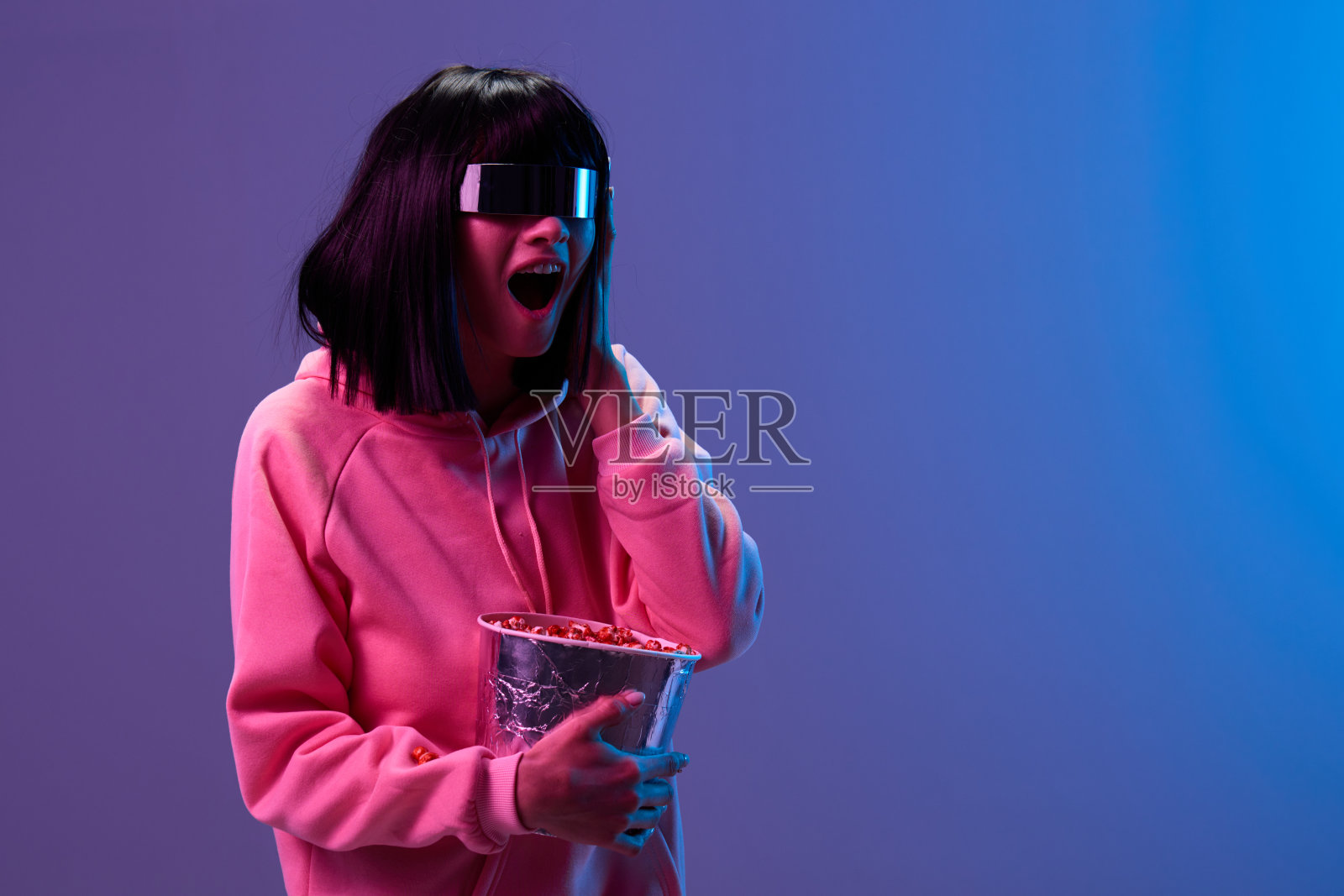 喜出望外的兴奋漂亮的黑发女士在粉红色的连帽衫镜面太阳镜爆米花张开嘴触摸脸颊摆出孤立的蓝紫色光背景。霓虹灯派对电影院概念。本空间照片摄影图片