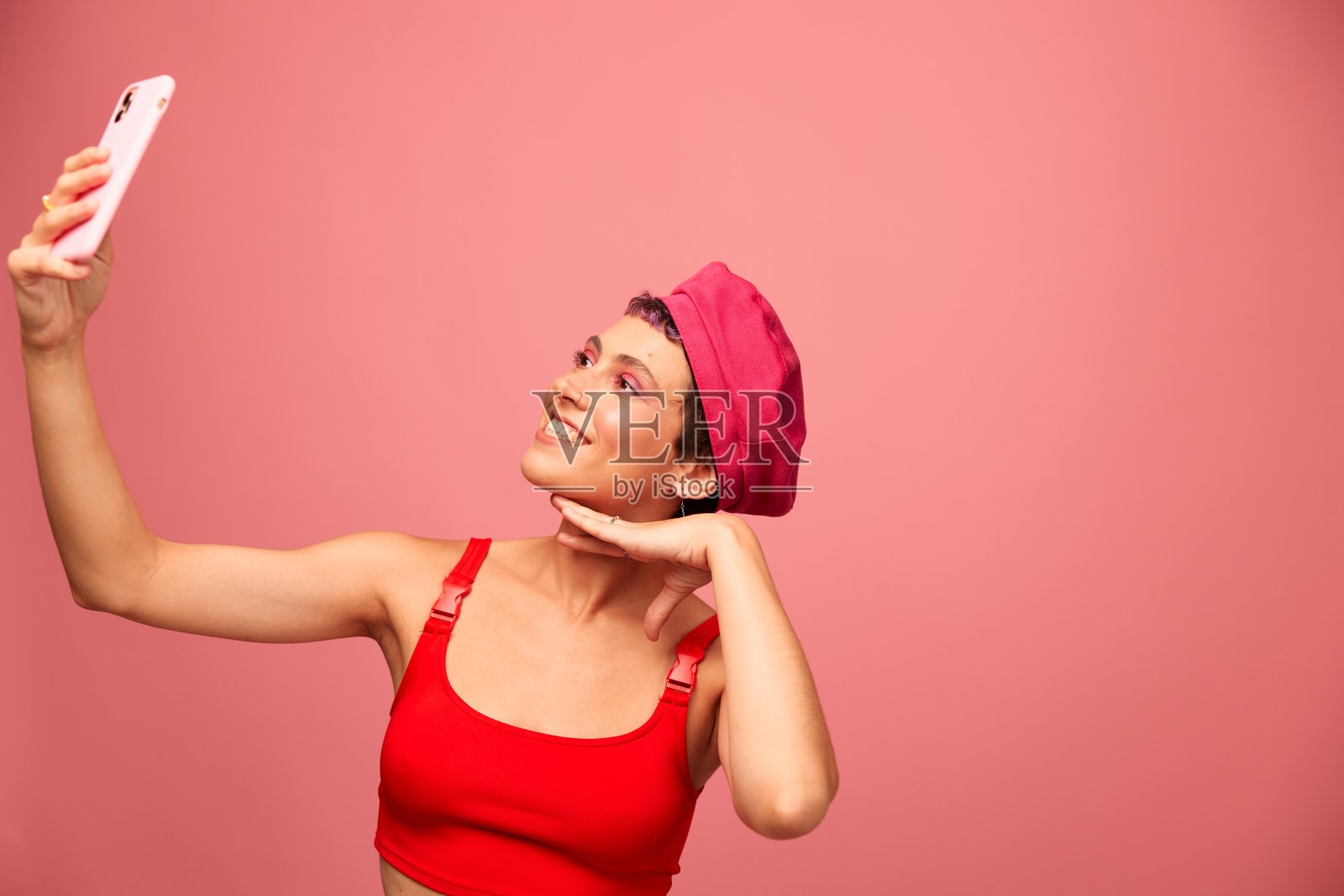 一位留着粉色头发和短发的年轻女博主用手机拍了一张自己的照片，她穿着时髦的衣服，戴着粉红色背景的单色帽子，脸上挂着微笑照片摄影图片