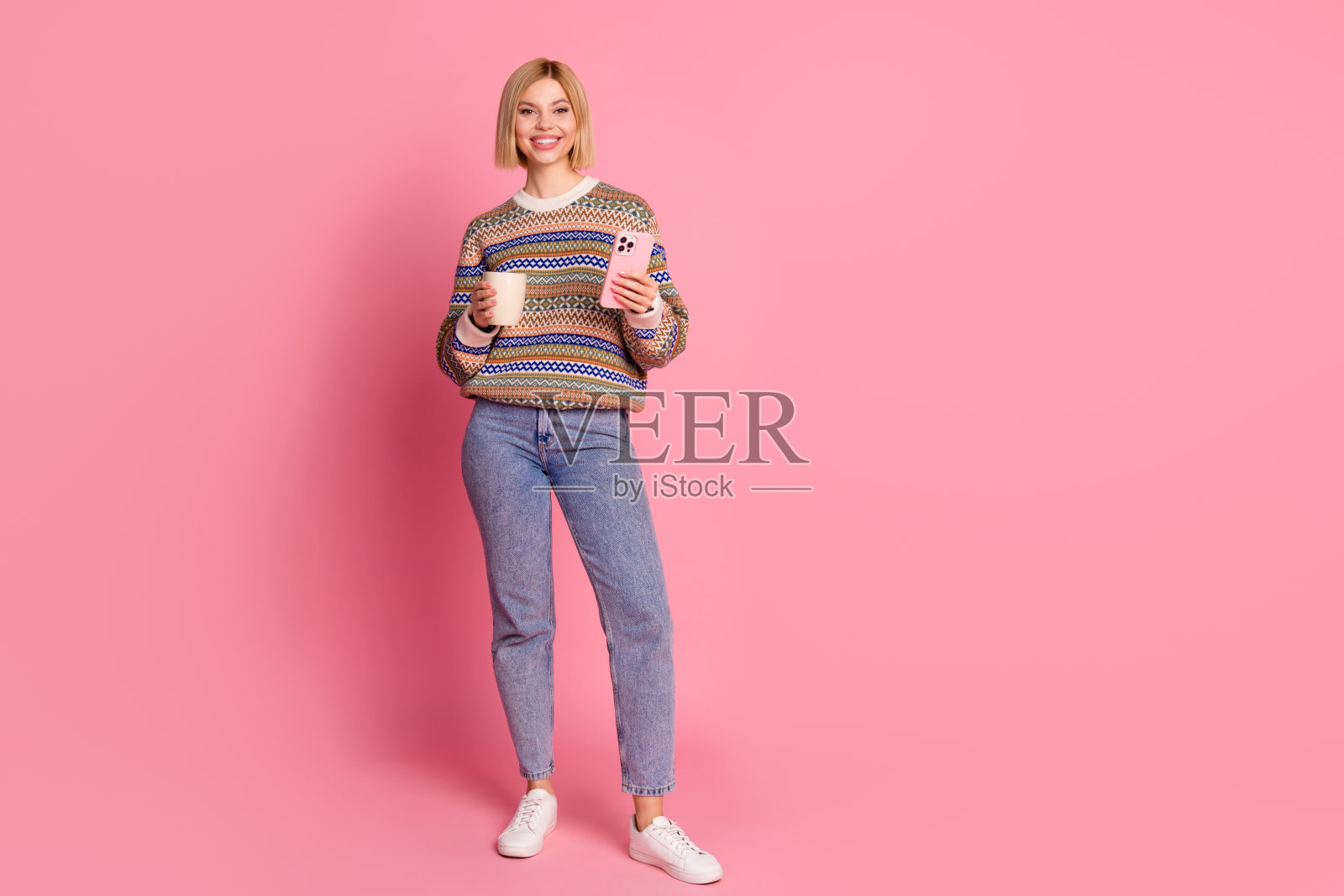 聪明可爱的女孩穿着印花毛衣牛仔裤的全身照片，拿着卡布奇诺杯，智能手机孤立在粉红色的背景上照片摄影图片