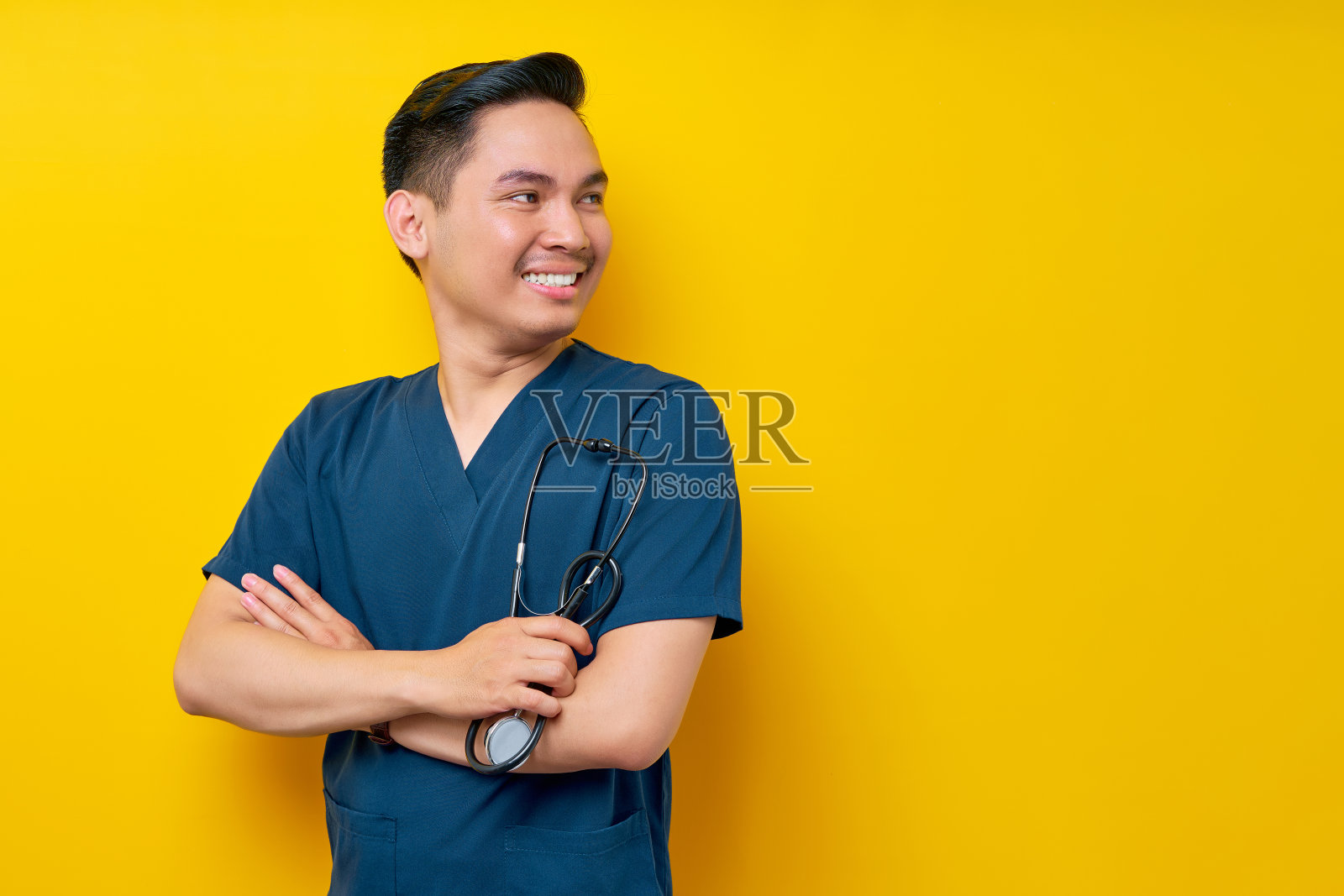 穿着蓝色制服的年轻专业亚洲男性医生或护士自信地站着，手抱听诊器，双手交叉，微笑友好地站在黄色背景上照片摄影图片