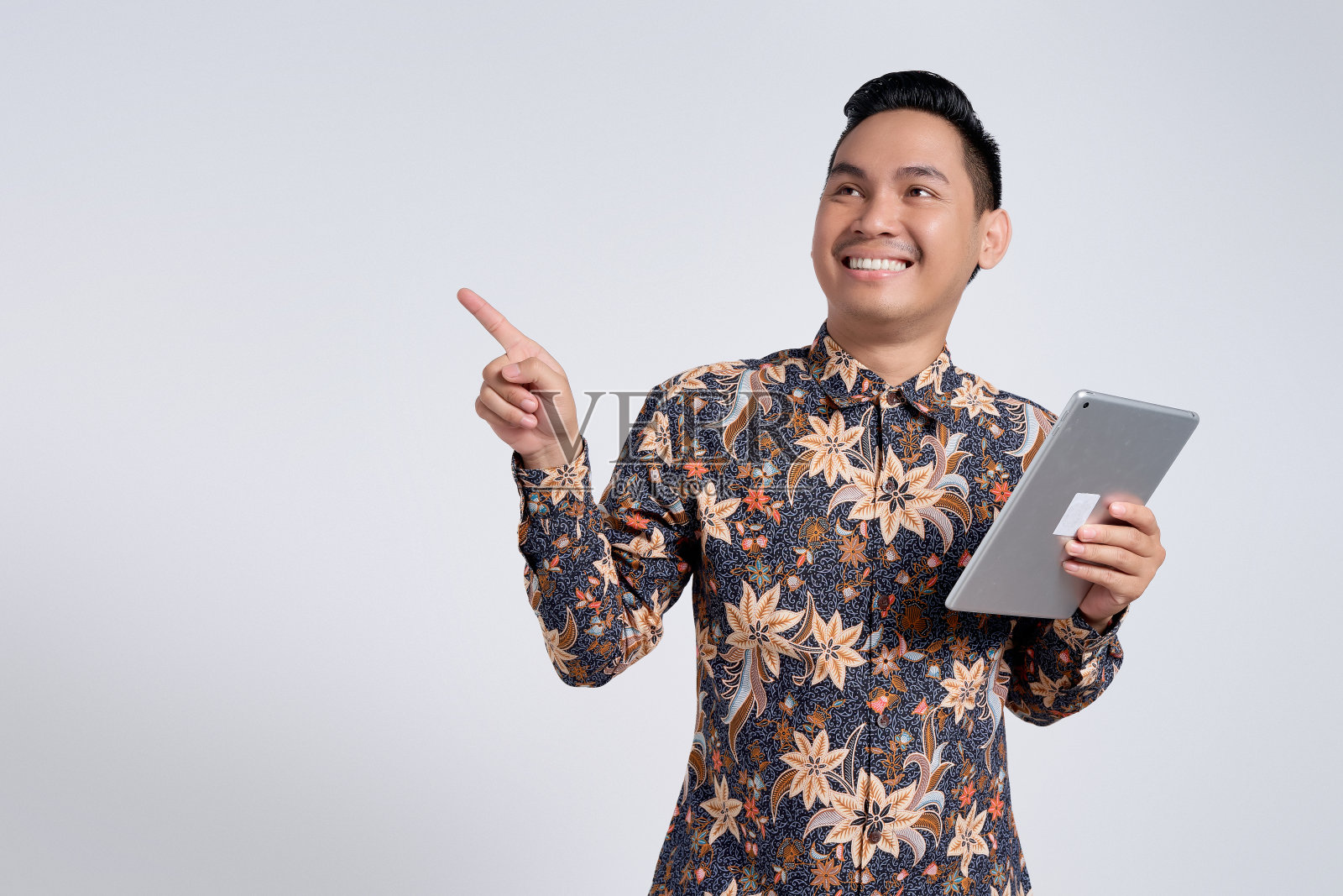 微笑的快乐英俊的年轻亚洲男子穿着蜡染衬衫拿着数码平板电脑，用手指指着白色背景上孤立的空白。人们的生活方式理念照片摄影图片