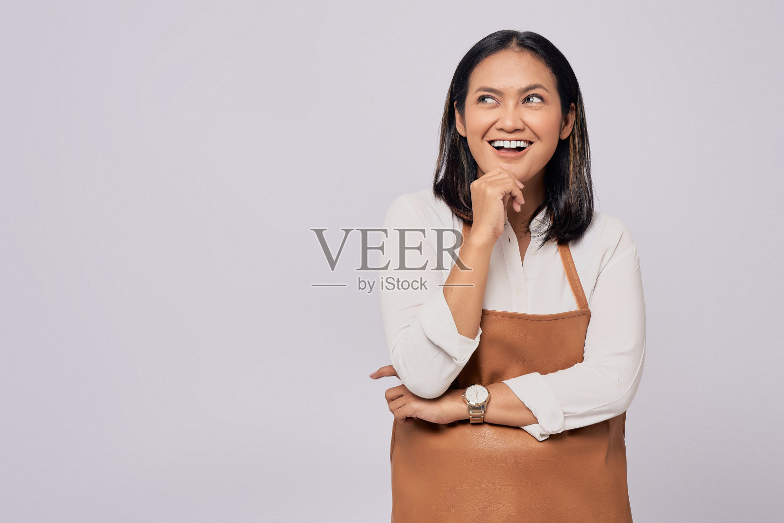 穿着棕色围裙在咖啡店工作的年轻亚洲女咖啡师酒吧服务员微笑着看向一边，触摸着白色背景上孤立的下巴。小型创业公司照片摄影图片