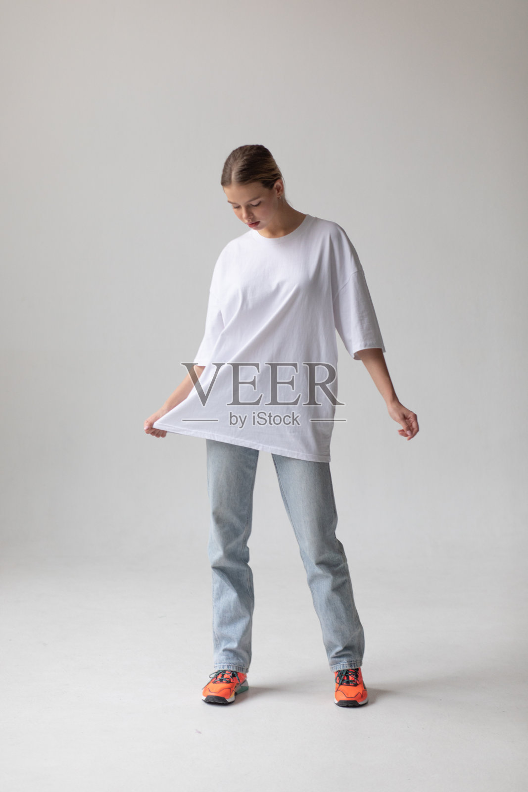 漂亮的金发女孩穿着白色的大号t恤和蓝色牛仔裤，在灰色的背景上摆姿势照片摄影图片