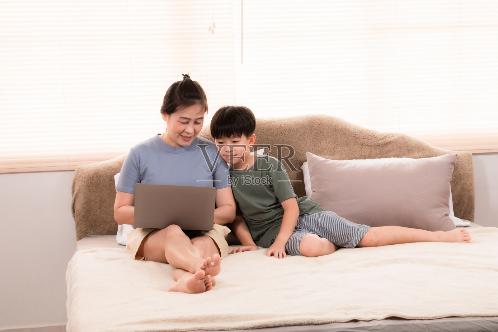 亚洲快乐的妈妈在床上用笔记本电脑工作，而儿子坐在旁边想和妈妈一起玩，单身妈妈一边工作一边抚养男孩。孩子乞求她的关注。照片摄影图片