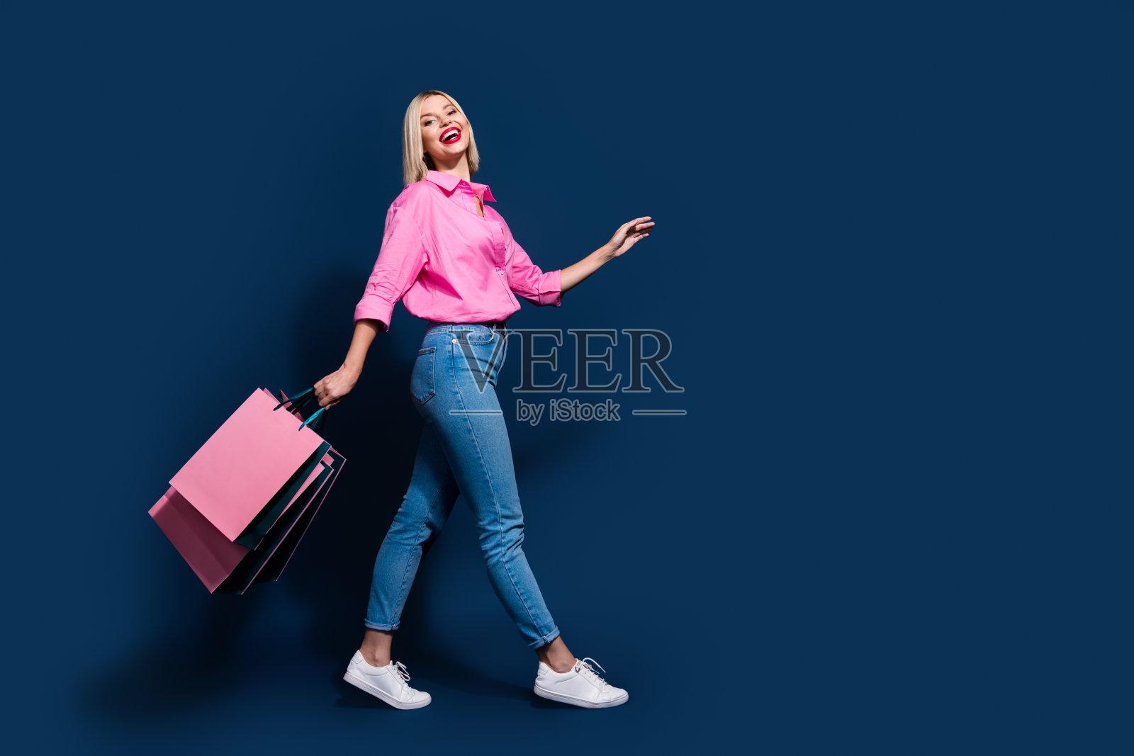可爱的年轻女士的全身照片，兴奋地走着，拿着购物袋，穿着时髦的粉红色衣服，与深蓝色的背景隔绝照片摄影图片