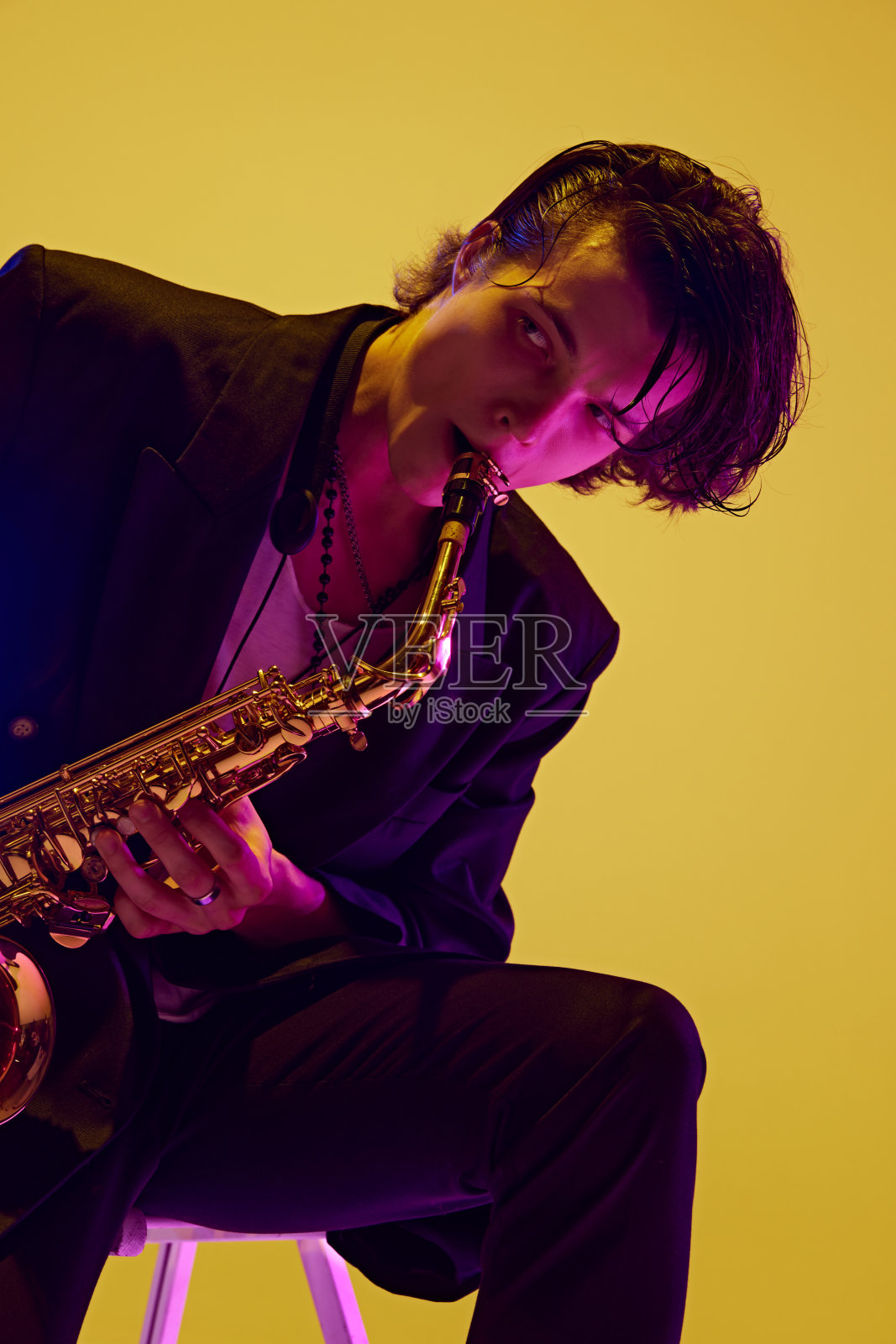 身穿深色西装的年轻迷人男子在霓虹灯下演奏萨克斯风，背景是充满活力的黄色工作室。照片摄影图片