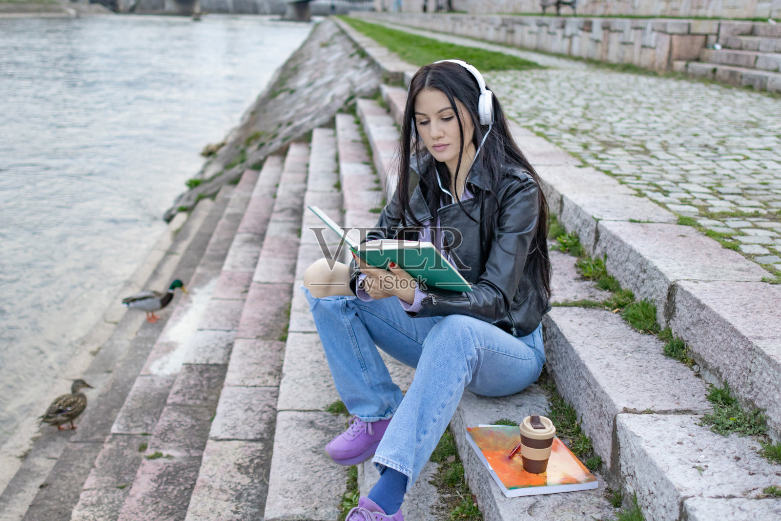 年轻漂亮的女大学生背着背包在河边玩。她戴着耳机，穿着皮夹克，等待着大学课程的开始。照片摄影图片