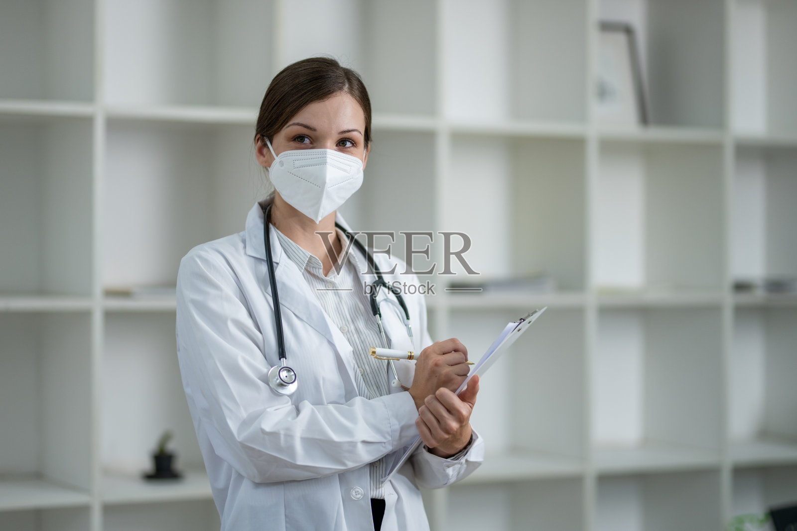 在现代诊所里，一位戴着防护口罩、手持病人病历的专业医护人员。照片摄影图片