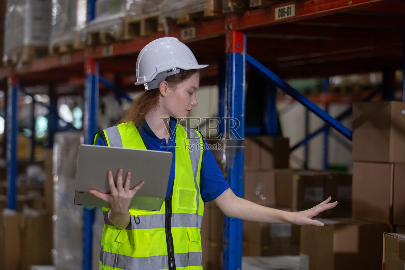 女工穿着安全制服，戴着安全帽，用笔记本电脑检查仓库货架上的货物。女工在工厂验货。照片摄影图片