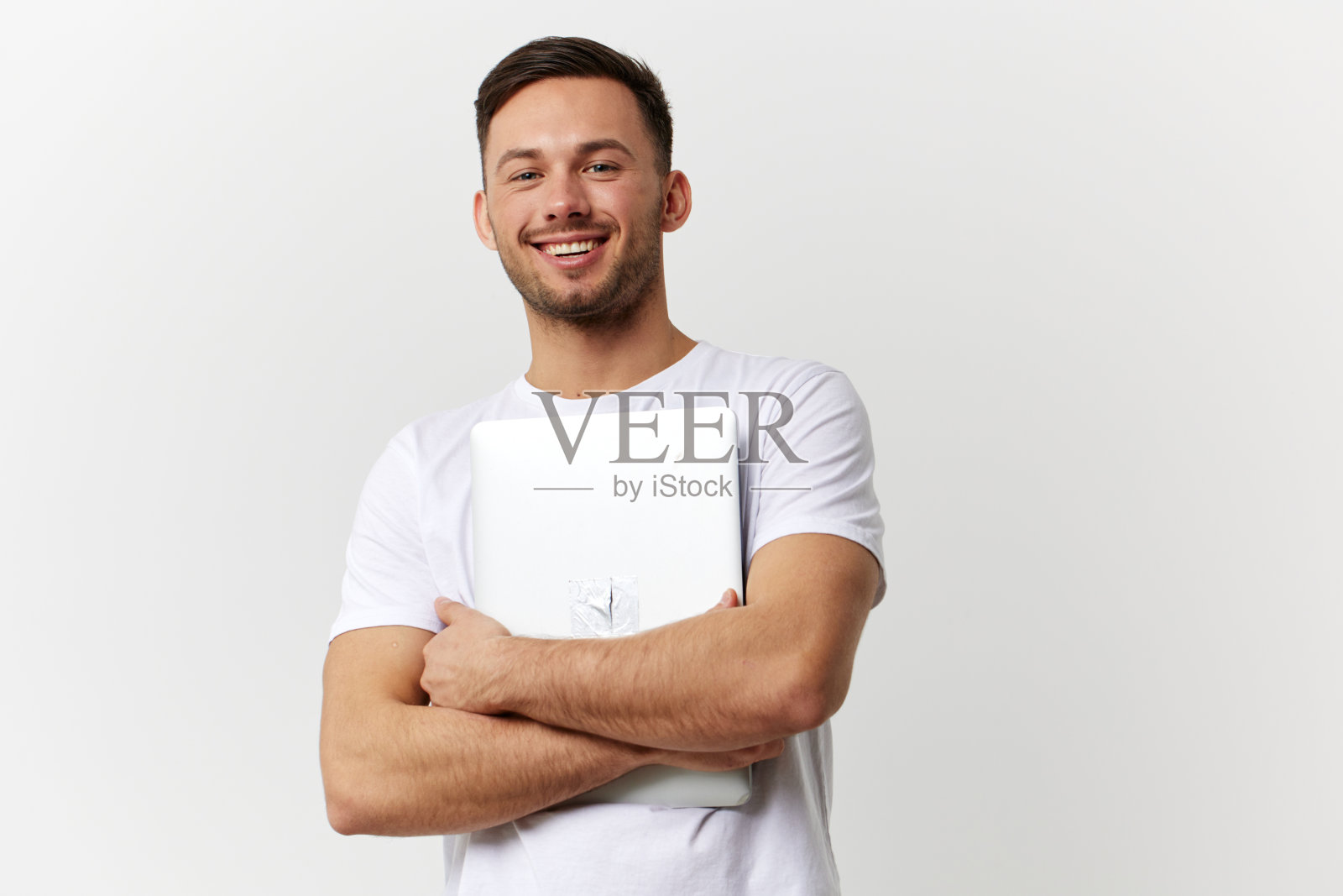 开朗、快乐、黝黑的英俊男子穿着基本的t恤，抱着笔记本电脑，在白色的工作室背景上摆出孤立的姿势。复制空间横幅模型。电子维修小玩意的生活理念照片摄影图片