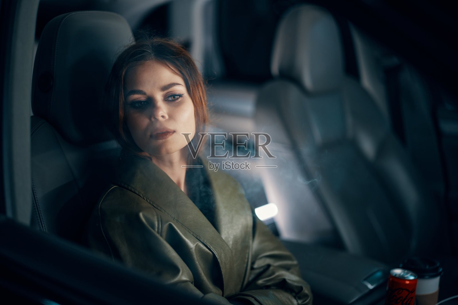 这是一幅近距离的横向肖像，一个穿着皮衣的时髦、奢华的女人晚上坐在一辆黑色汽车的副驾驶座位上，透过雾气蒙蒙的玻璃向一侧看照片摄影图片