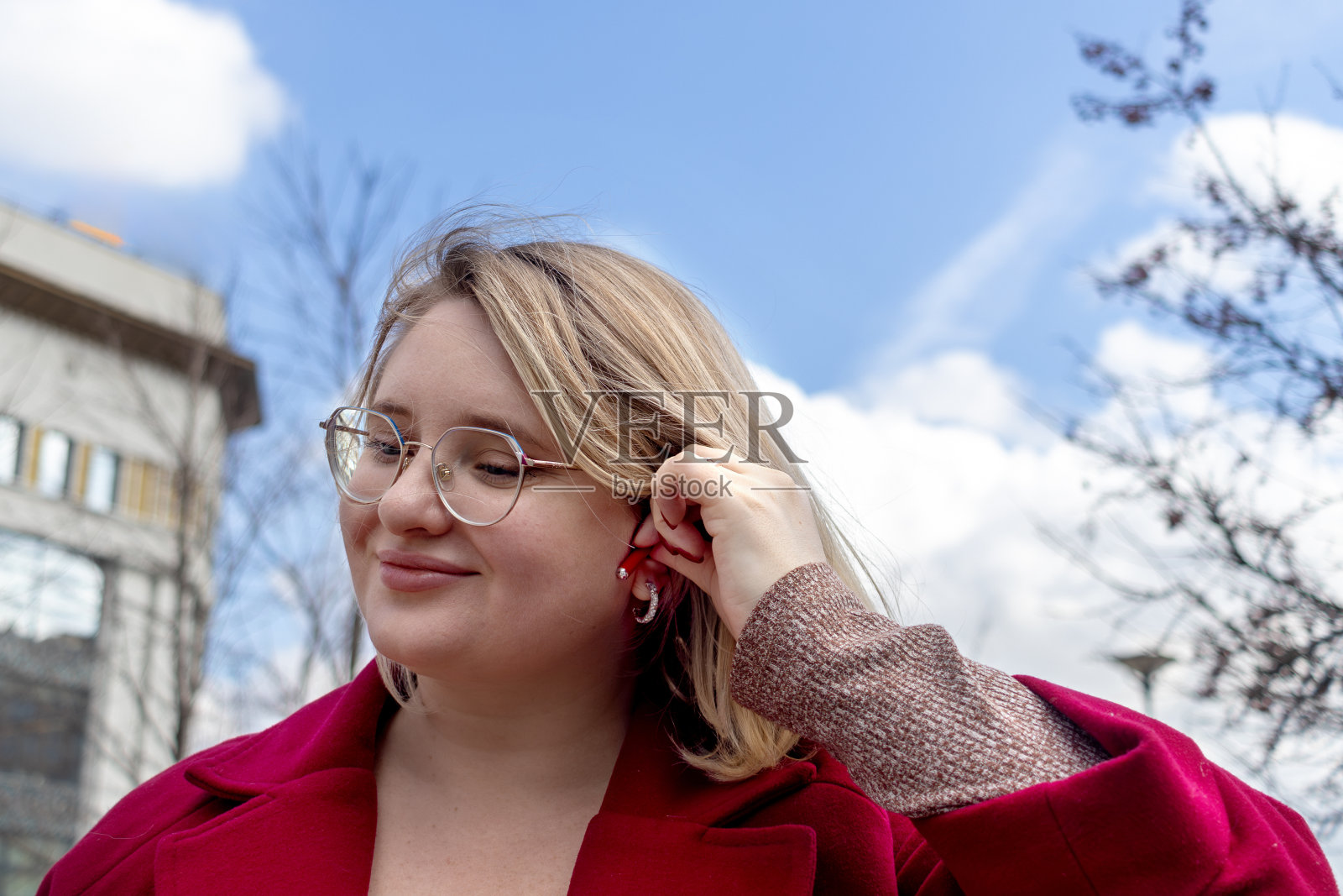 使用无线耳机。穿红色外套的大码年轻女子在城市里走着，耳朵里戴着耳机。特写镜头照片摄影图片