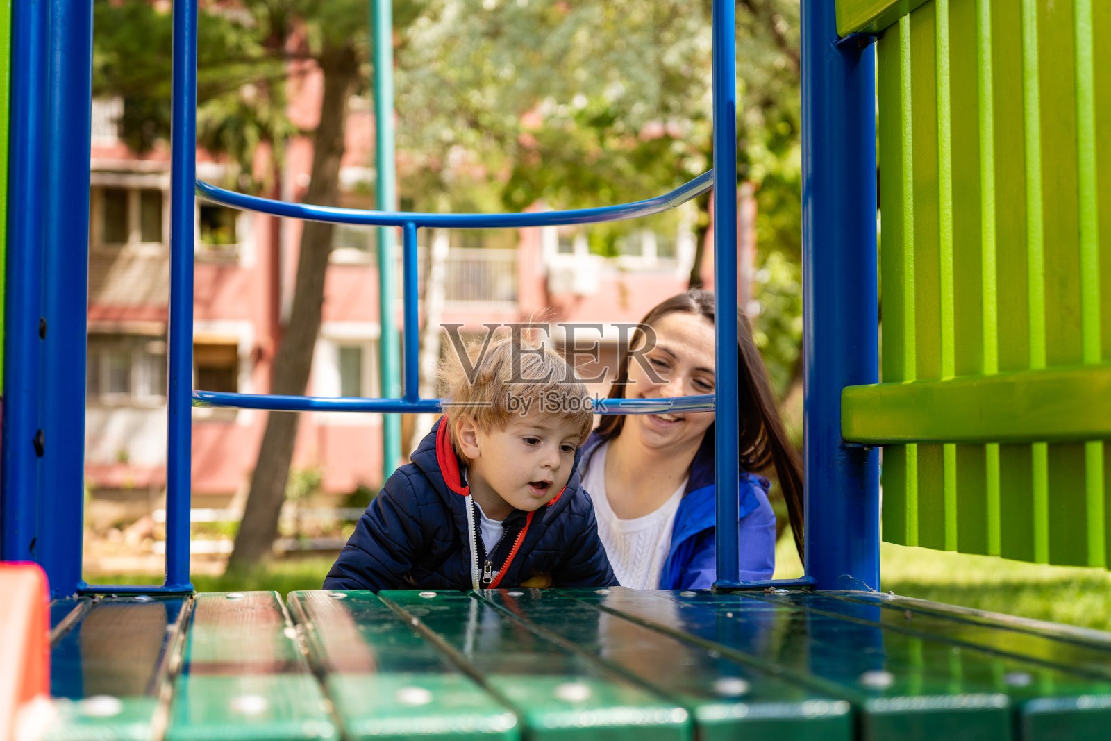一个快乐、强壮、健康的孩子正在儿童游乐场里攀爬，妈妈在一旁照看着他照片摄影图片