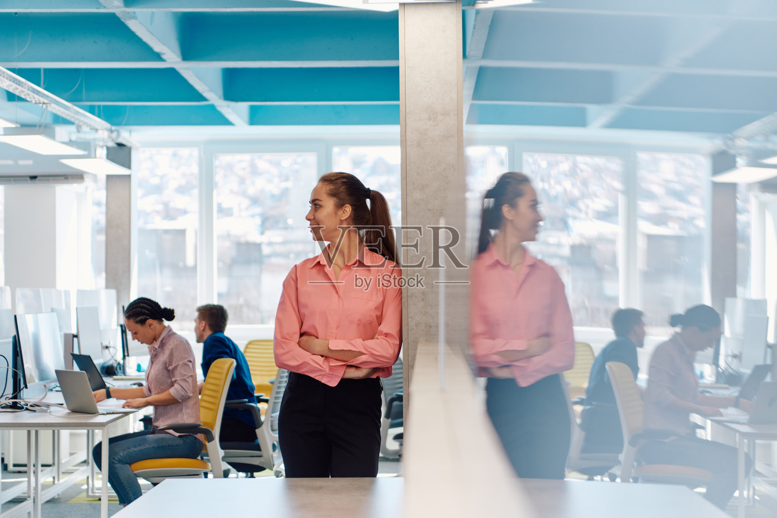 年轻微笑的商业女性在创意开放空间联合创业办公室的肖像。成功的女商人站在办公室拿着广告版面。在后台工作的同事照片摄影图片