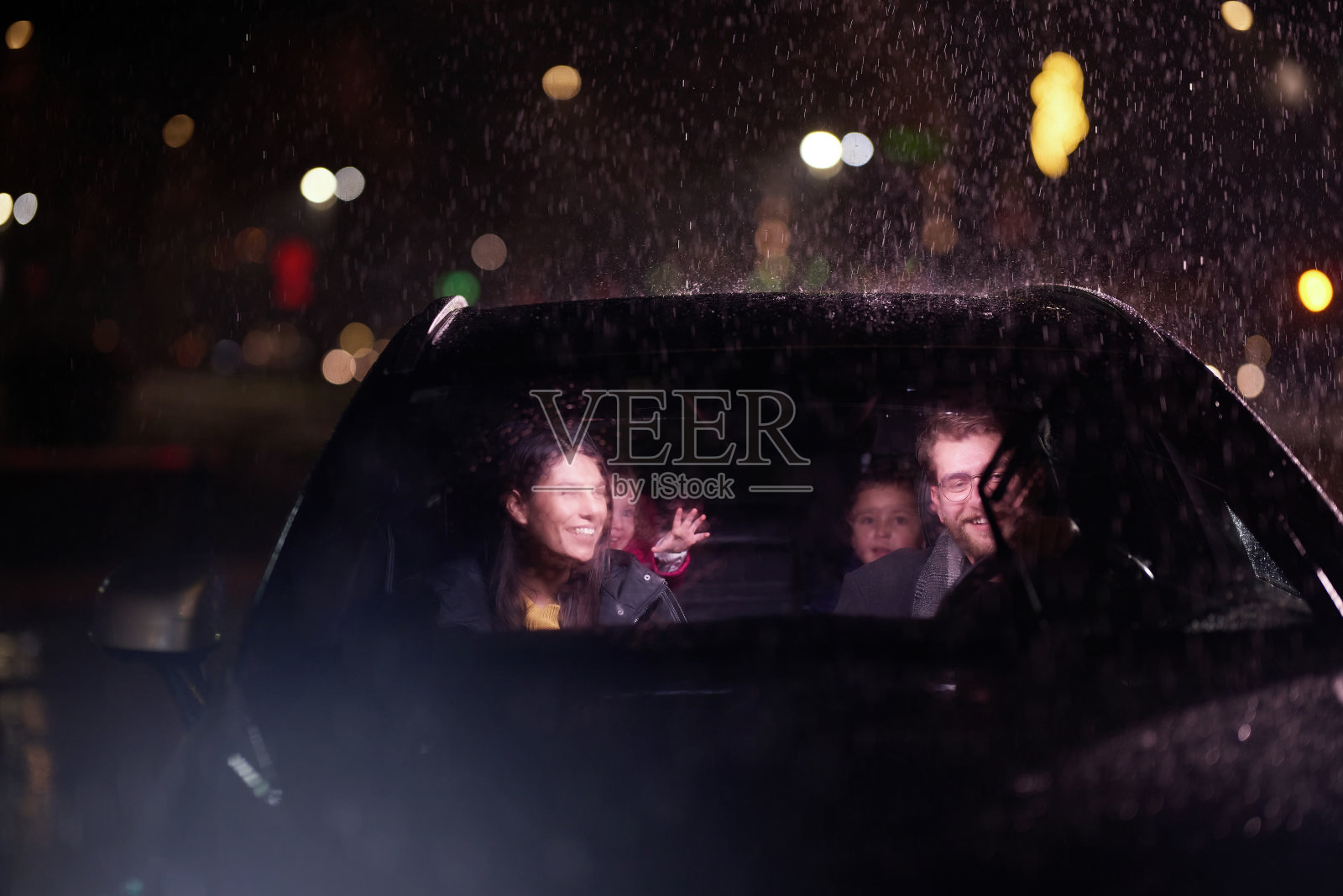 在夜晚的旅程中，一个幸福的家庭在雨天行驶的汽车里享受着欢乐的时刻，车头灯的光芒、笑声和亲情照亮了他们照片摄影图片