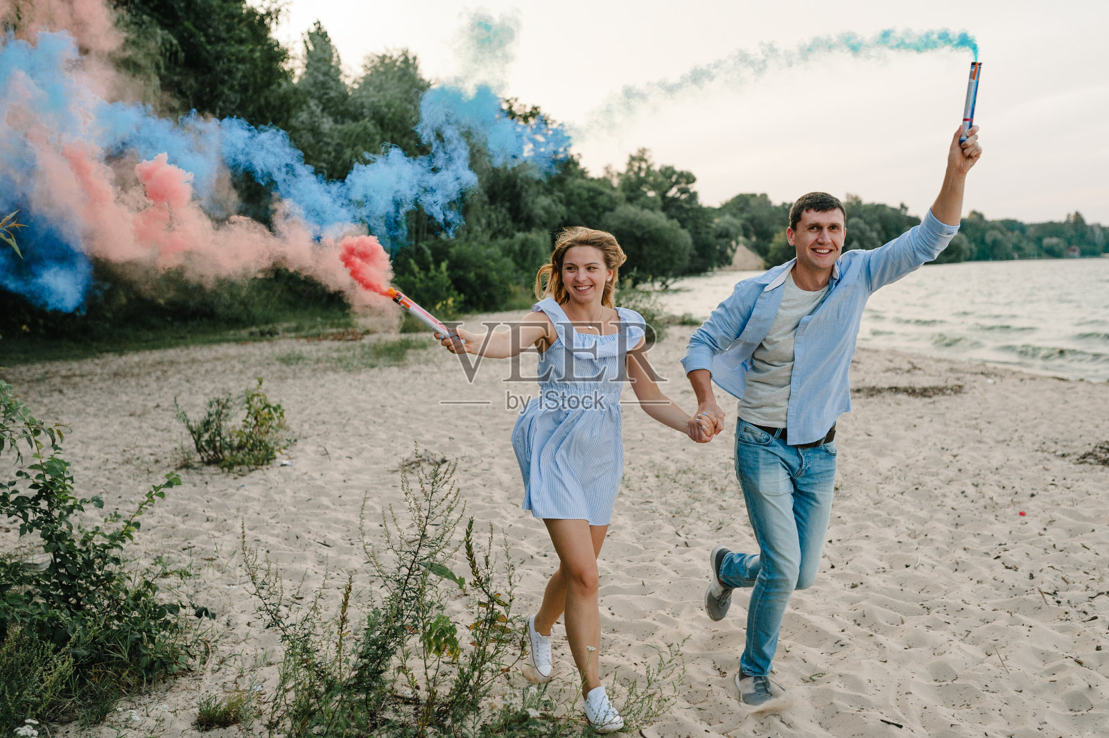幸福的已婚夫妇手牵手烟雾弹。男人和女人在沙滩上奔跑，拿着蓝色、红色、粉色的烟雾弹，看着海滩上的相机，玩得很开心。婴儿洗澡。双胞胎:男孩和女孩照片摄影图片