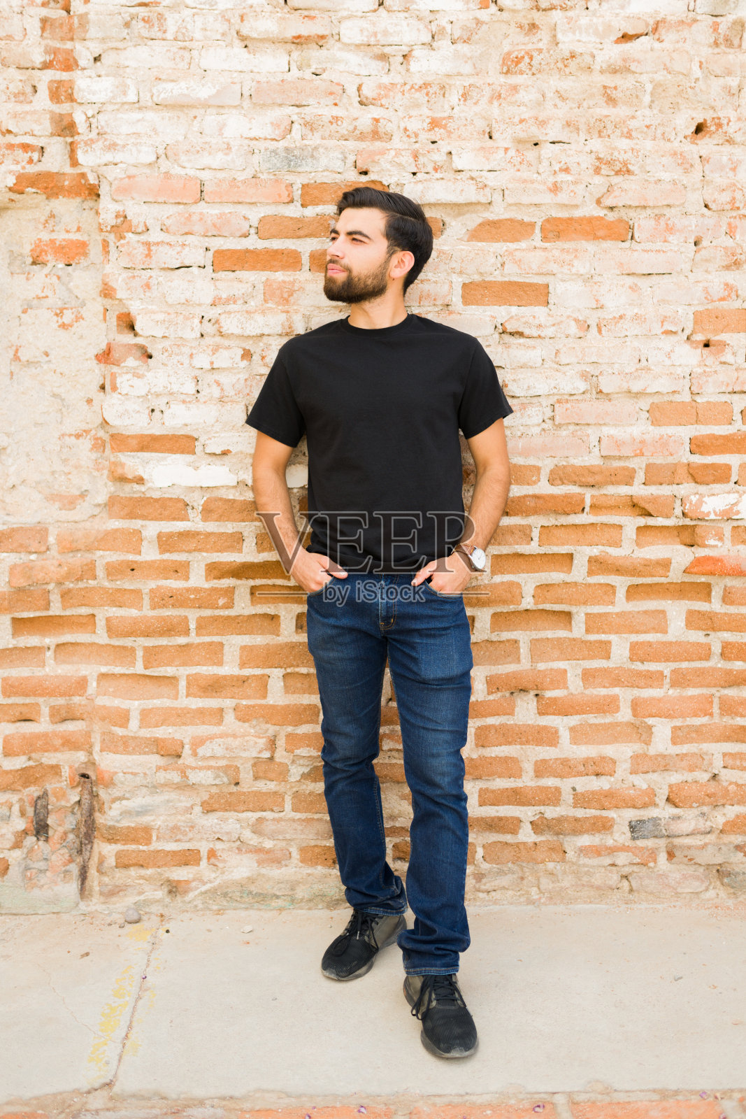 自信的年轻西班牙人穿着一件空白的黑色t恤进行模型展示，站在质朴的砖墙背景下照片摄影图片