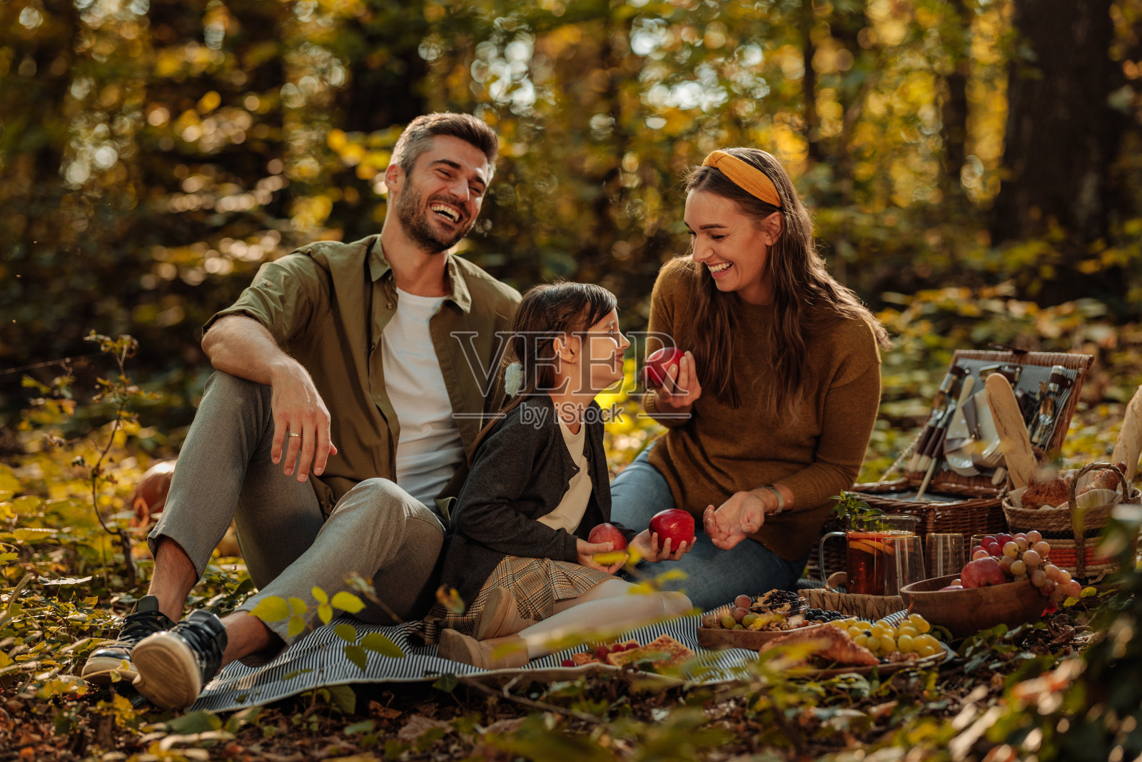 妈妈，爸爸和他们的小女儿坐在毯子上，在森林里拿着苹果照片摄影图片
