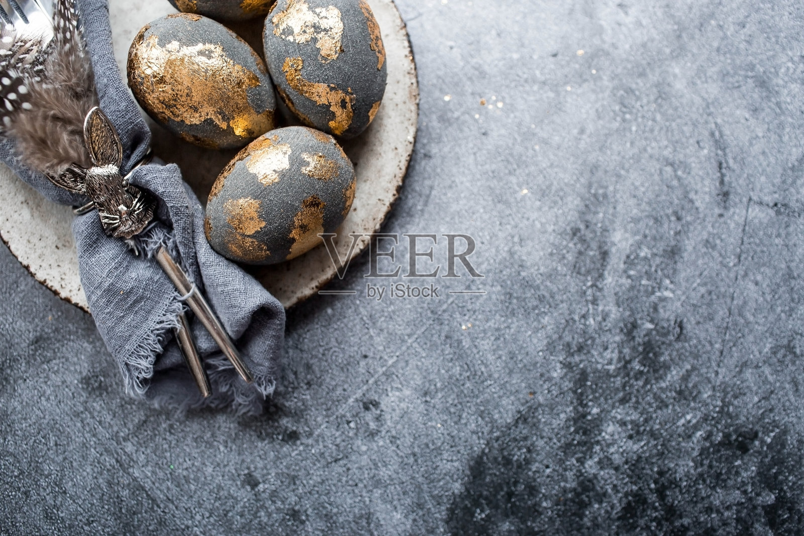 的横幅。表设置。一个时髦的混凝土盘子，餐巾上有一只兔子，镀金的复活节彩蛋，灰色背景上的羽毛。咖啡馆和餐馆的复活节快乐假期的概念。照片摄影图片