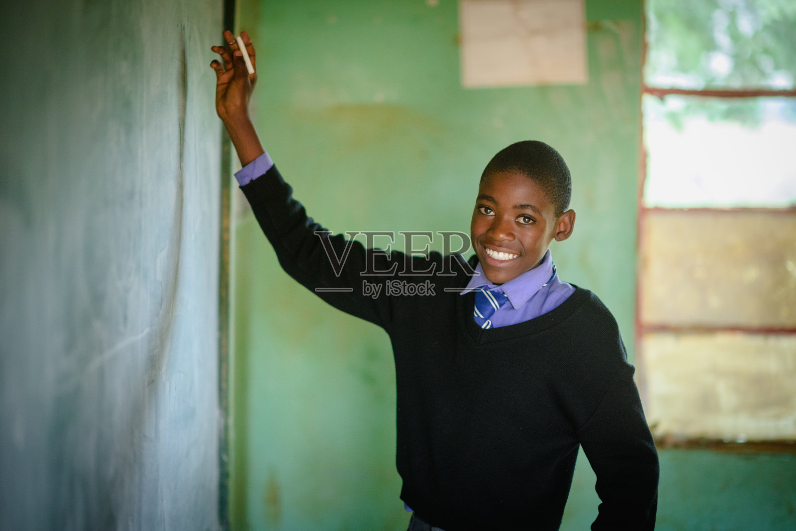 在一所乡村学校的教室里，穿着校服的非洲男生手持粉笔在黑板前微笑照片摄影图片