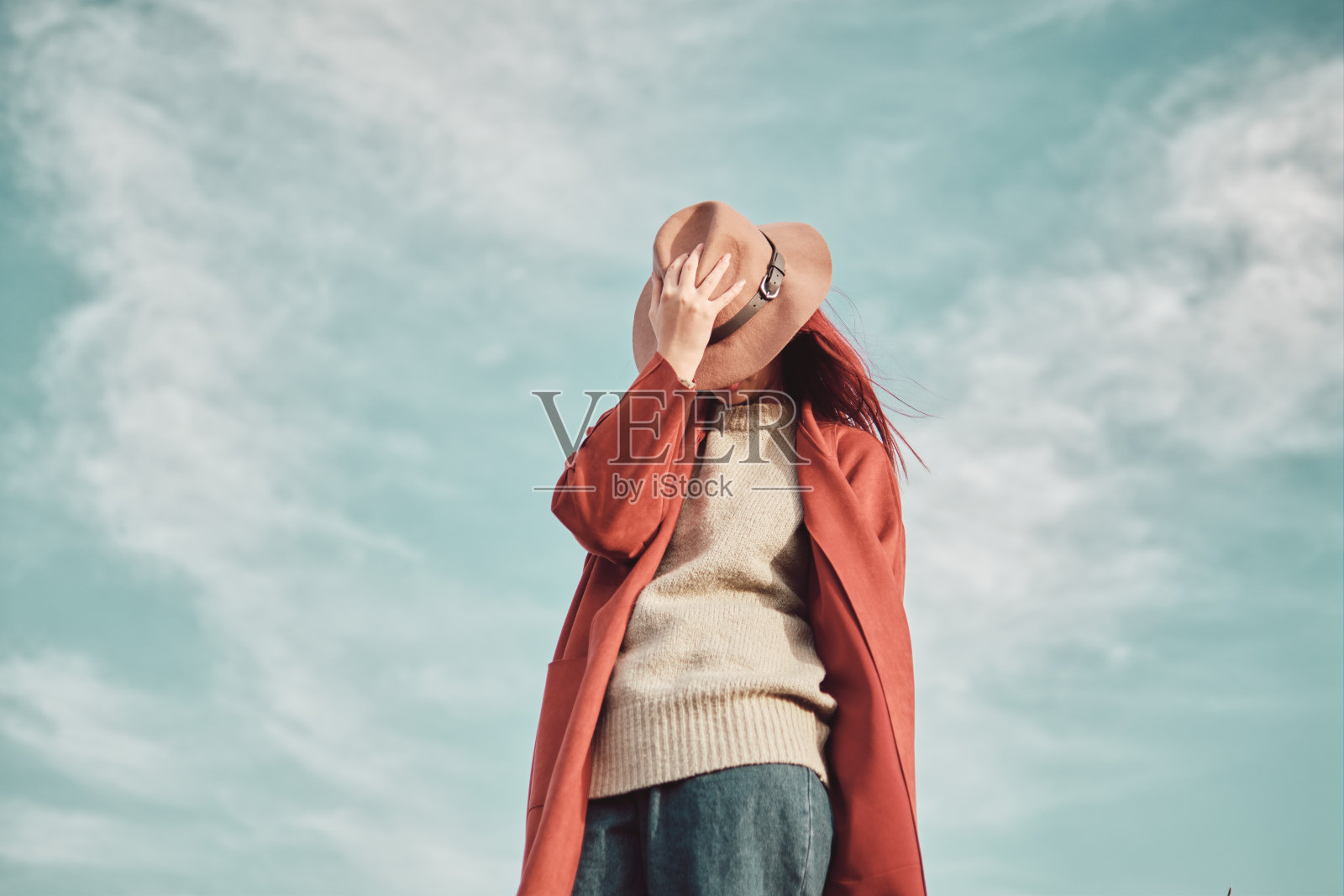 穿着红色外套的少女站在天空的衬托下，用帽子遮住脸。低角度视图。照片摄影图片