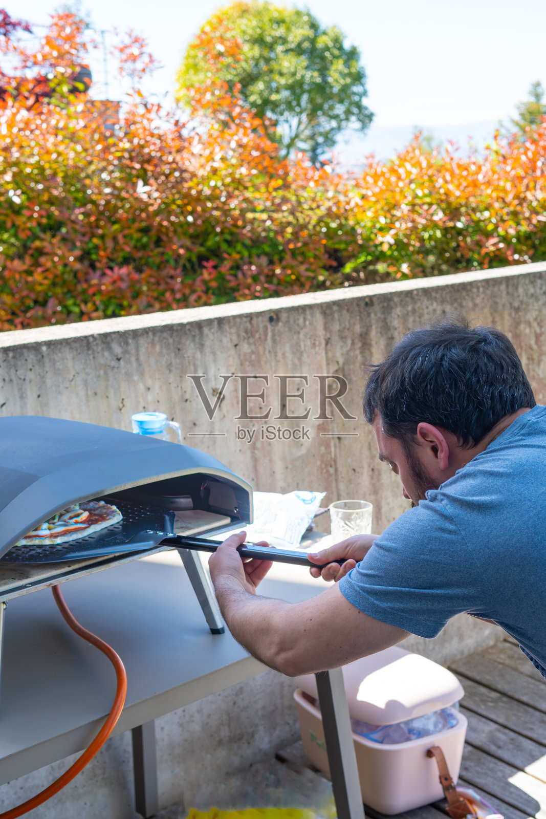 一个全神贯注的男人把自制的披萨铲进了他家露台上的煤气炉。树木背景照片摄影图片