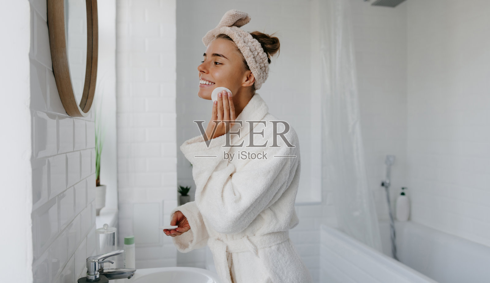 穿着浴袍的年轻美女站在浴室里用化妆棉洗脸照片摄影图片