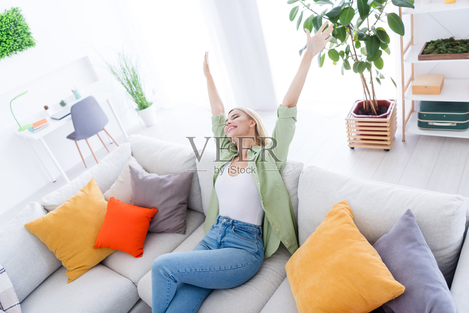 照片中的年轻开朗的金发无忧无虑的女人举起手臂，寒冷的周末放松地躺在客厅舒适的沙发上照片摄影图片