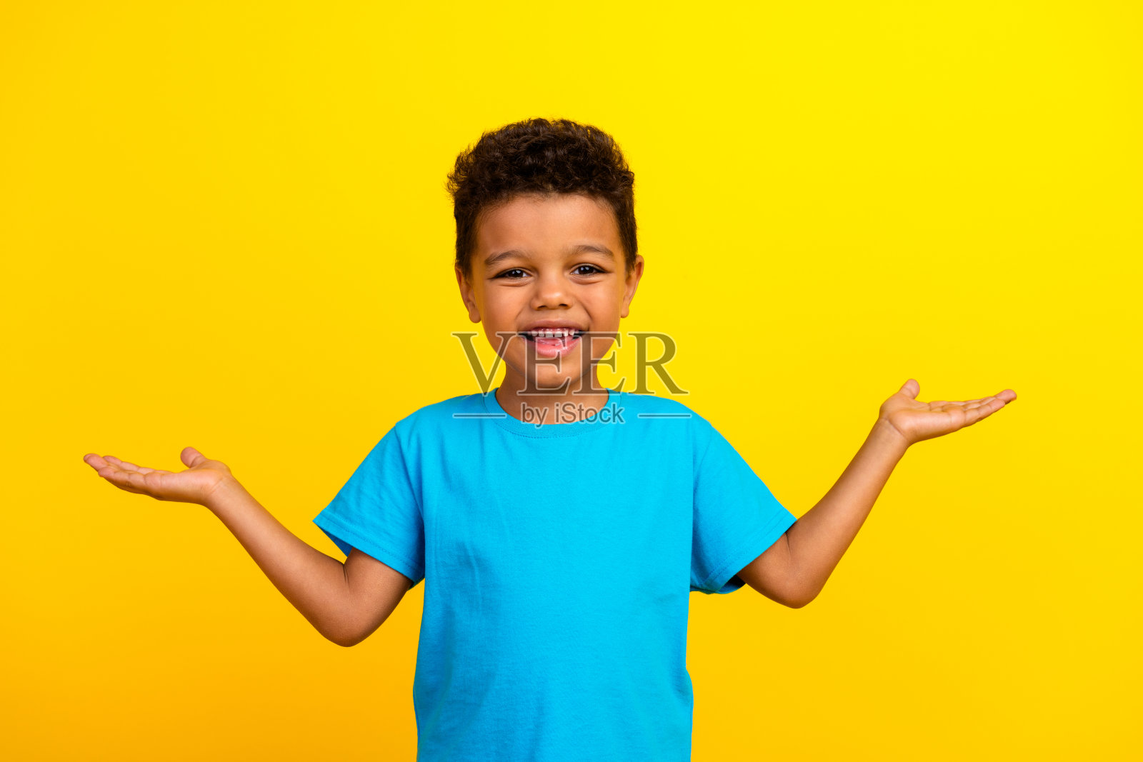 可爱的青春期前男孩的肖像，留着非洲式发型，穿着蓝色t恤，手掌比较产品，空白的空间孤立在黄色背景上照片摄影图片