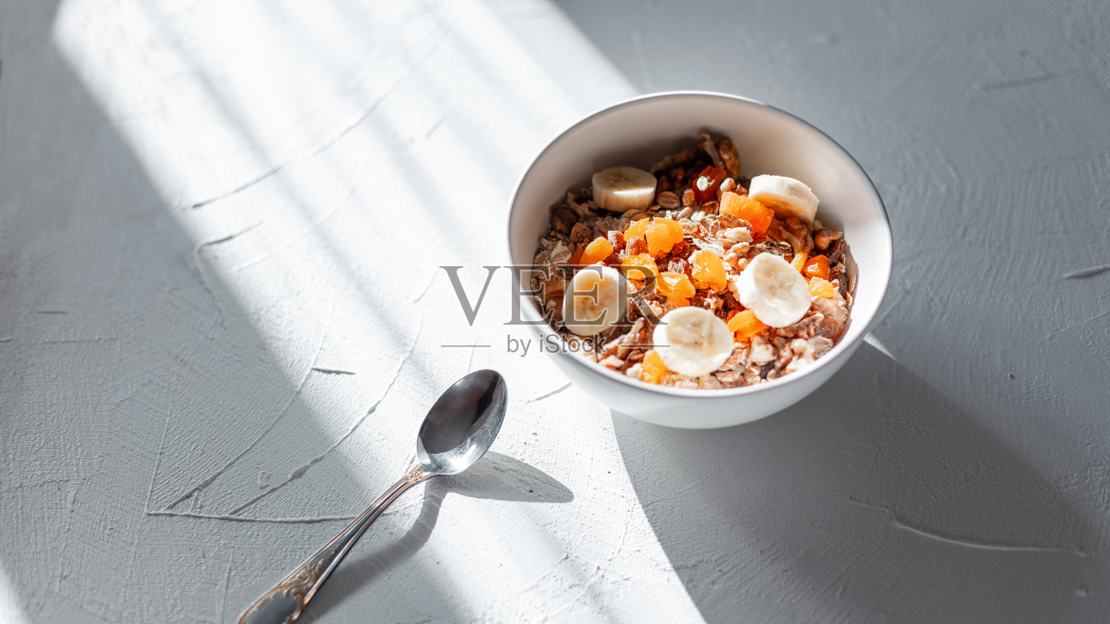 清晨日出时，白色的桌子上摆着美味的纯素早餐，燕麦片、杏干、坚果和香蕉装在一个白色的碗里。健康生食概念。极简主义照片摄影图片