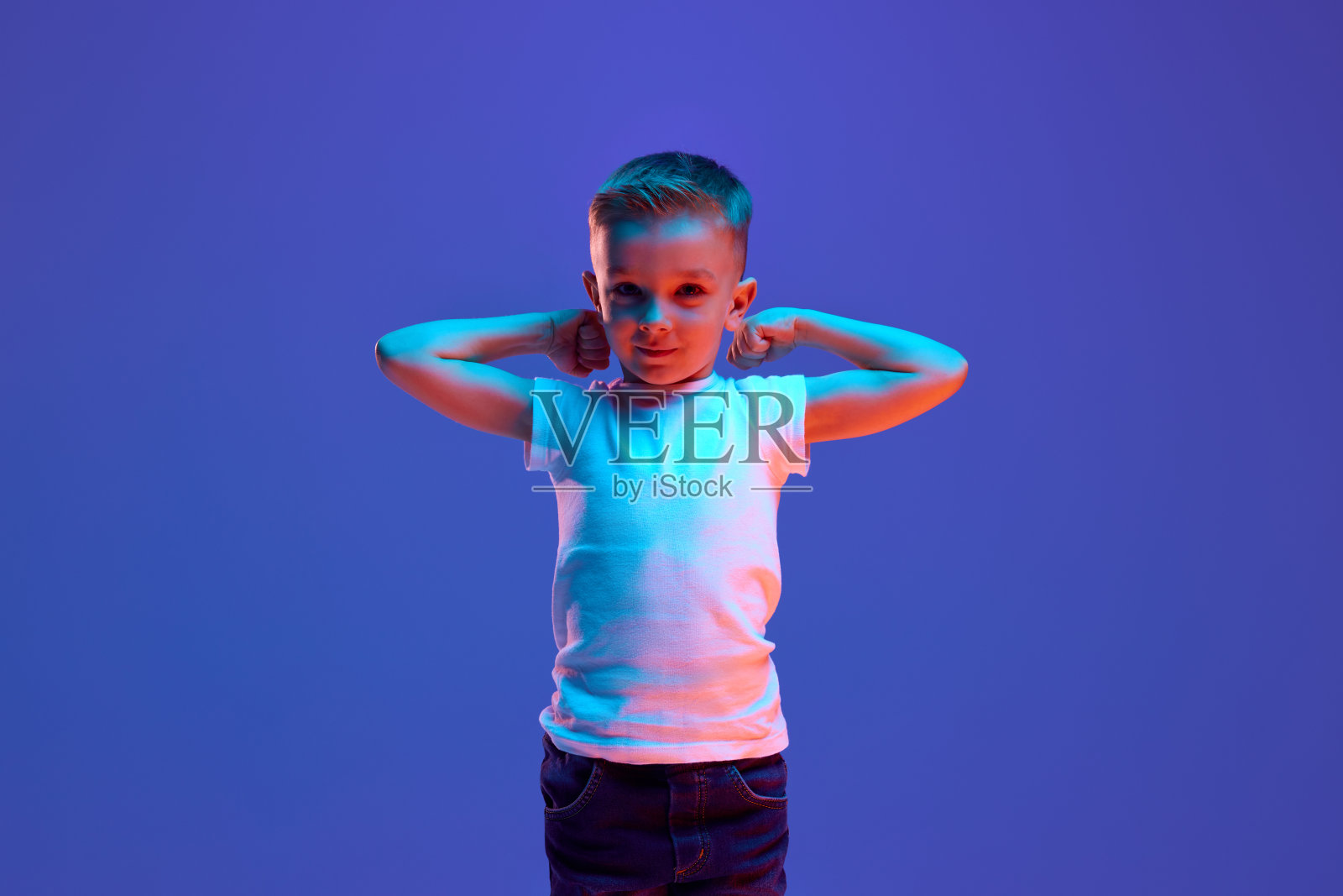 小男孩的肖像，在蓝色背景的混合霓虹灯下，男孩举起双手，展示他的力量，顽皮地看着相机。照片摄影图片