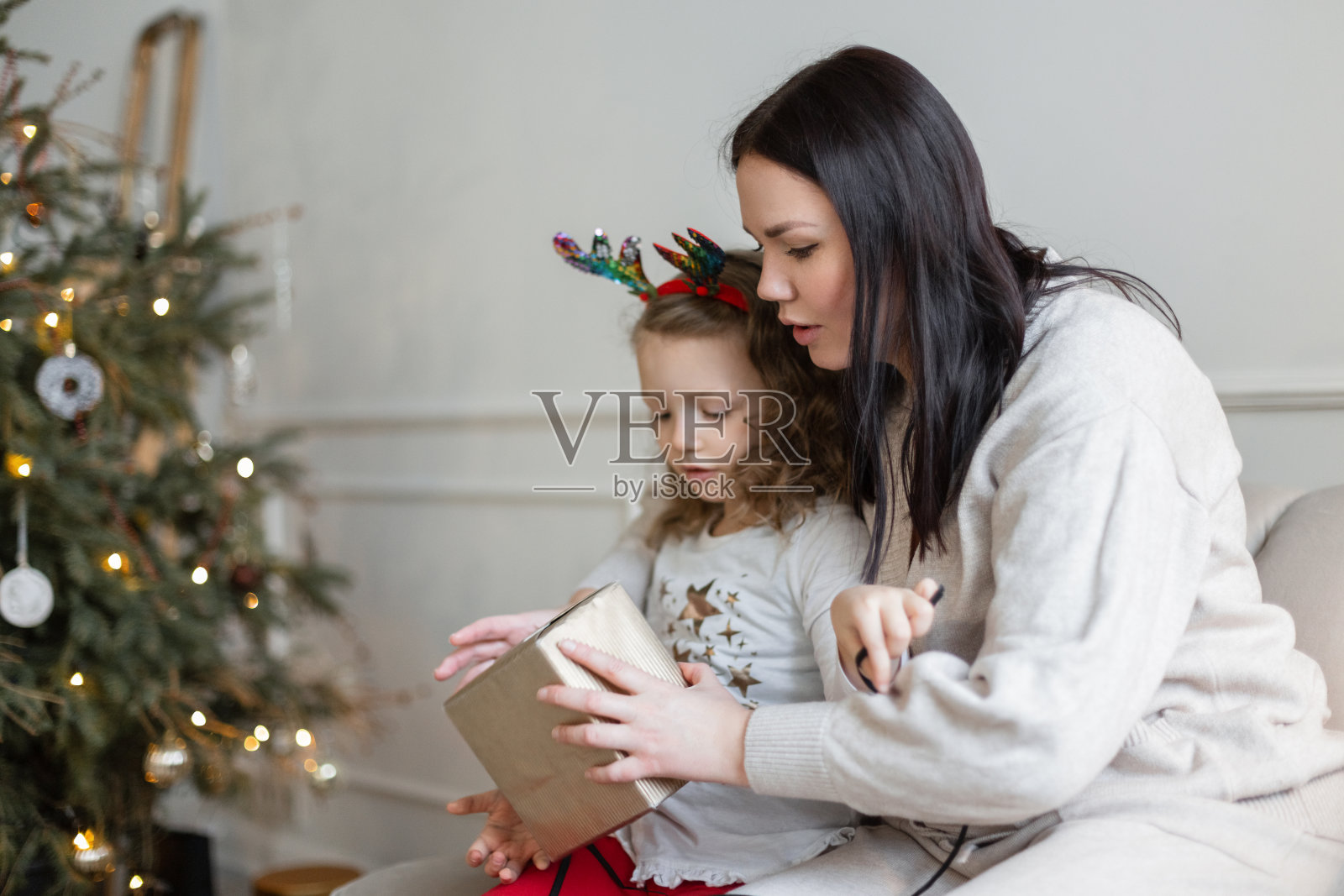 平安夜，穿着时髦衣服的妈妈和女儿坐在圣诞树旁的沙发上拆礼物照片摄影图片