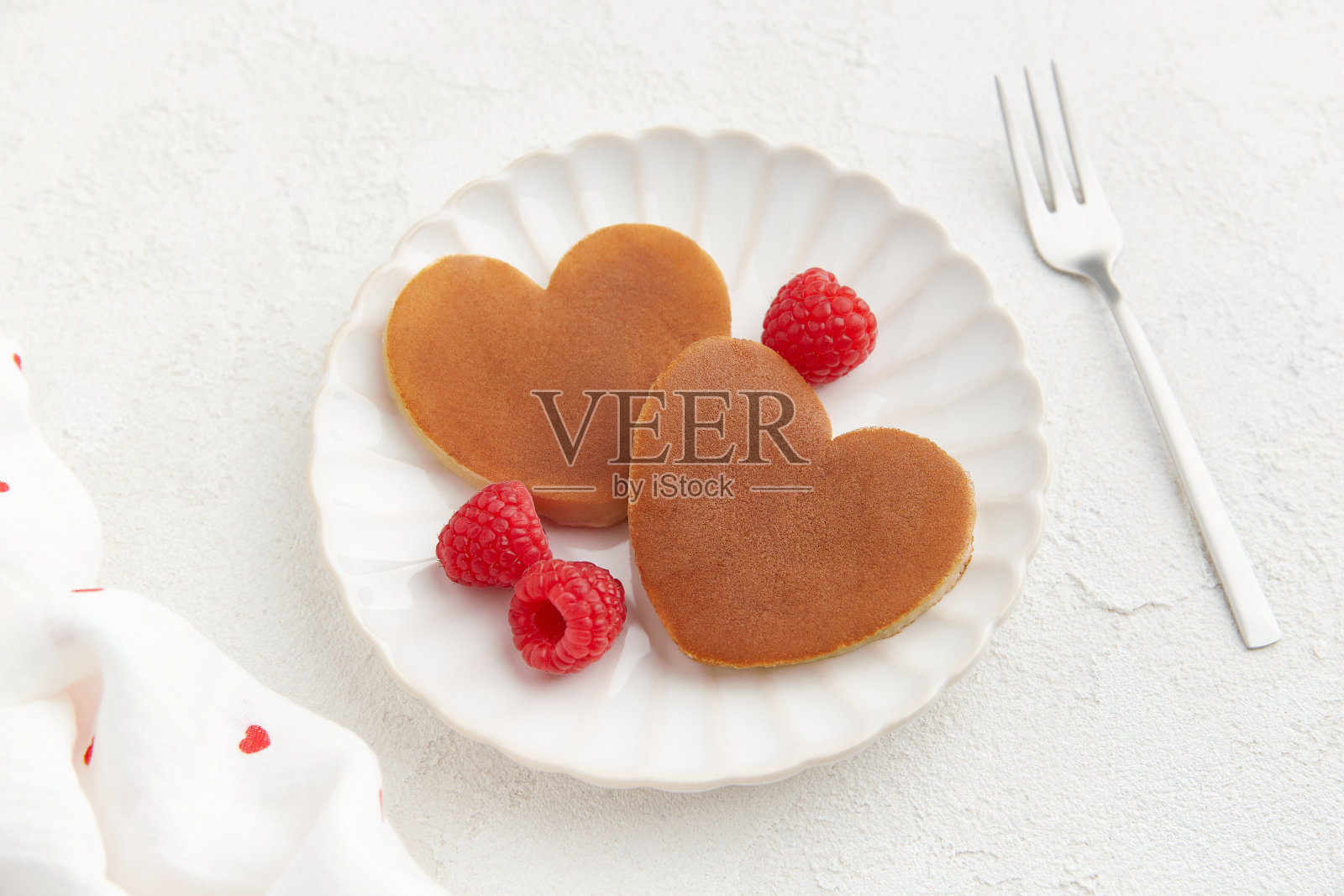 两个心形的煎饼，上面有覆盆子和一把叉子，放在白色的盘子上，白色的背景照片摄影图片
