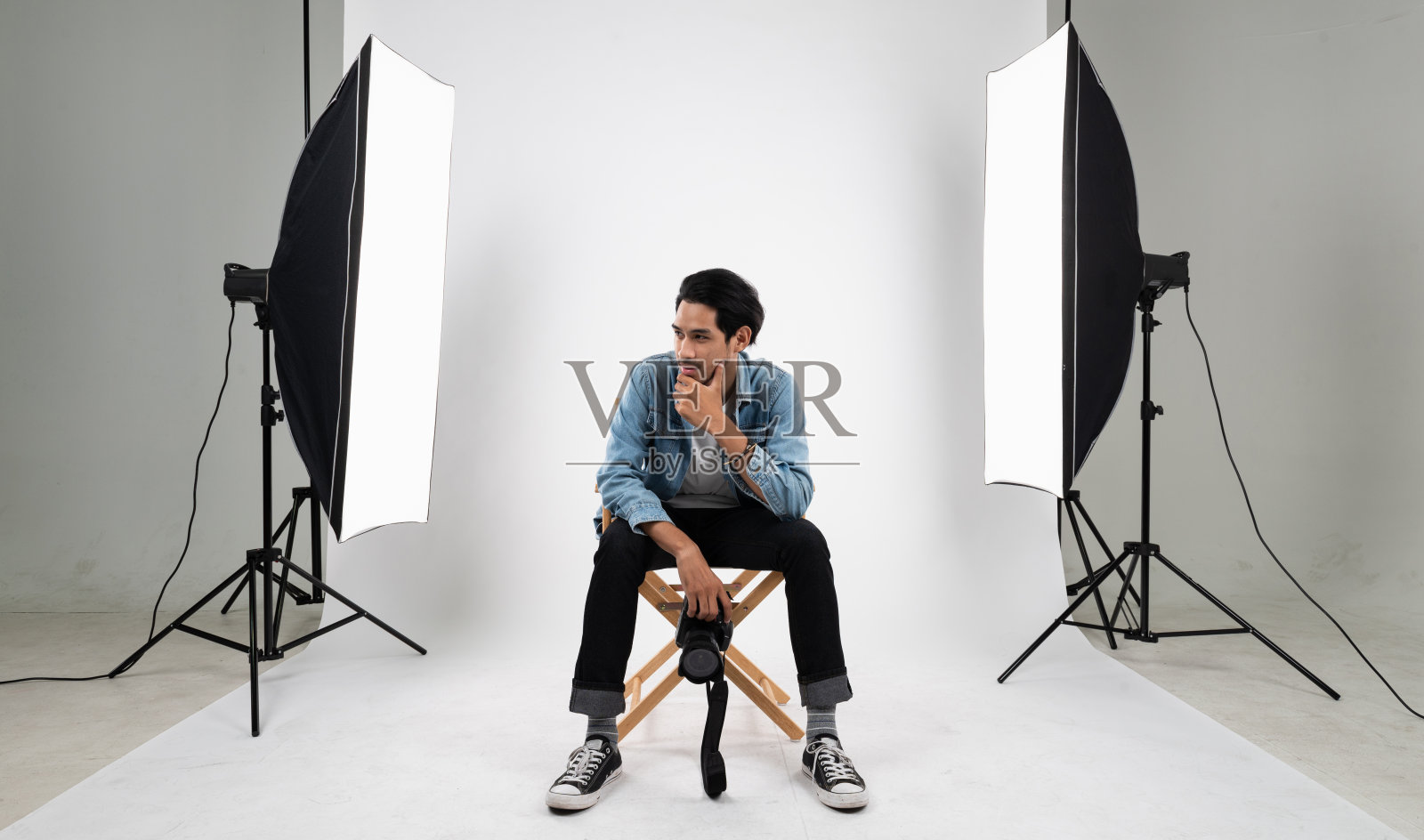 年轻聪明的亚洲专业摄影师的肖像照片坐在白色的照片背景室内摄影棚与一套频闪闪光灯和柔光箱准备产品和模型拍摄。照片摄影图片