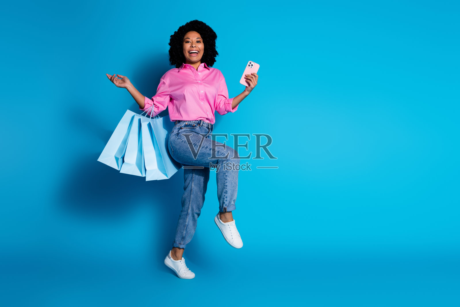 漂亮的年轻女子手持购物袋智能手机空的空间穿着粉红色的衬衫孤立在蓝色的背景照片摄影图片