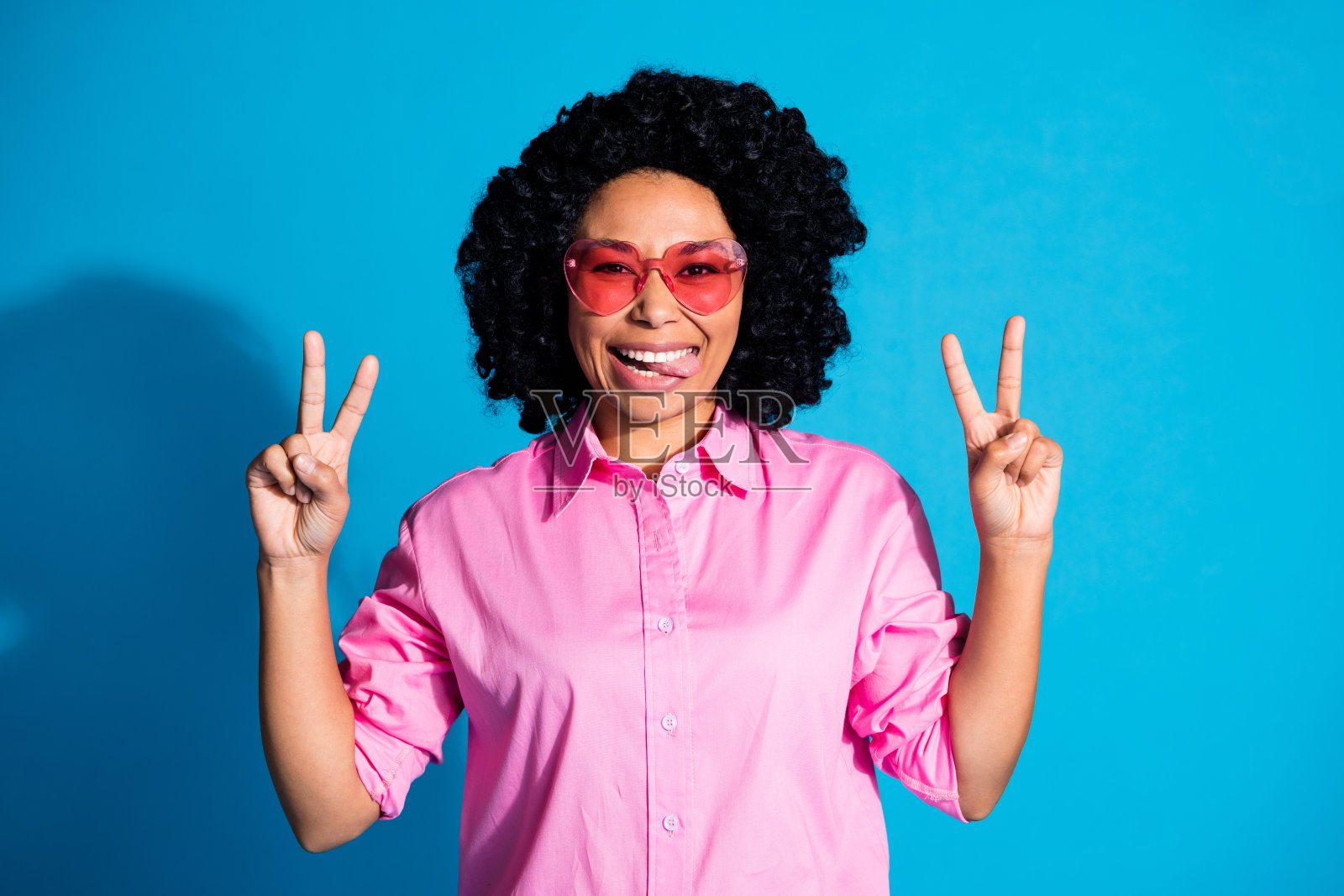 漂亮的年轻女子的肖像，露出舌头的v字，穿着粉红色的衬衫，孤立在蓝色的背景照片摄影图片