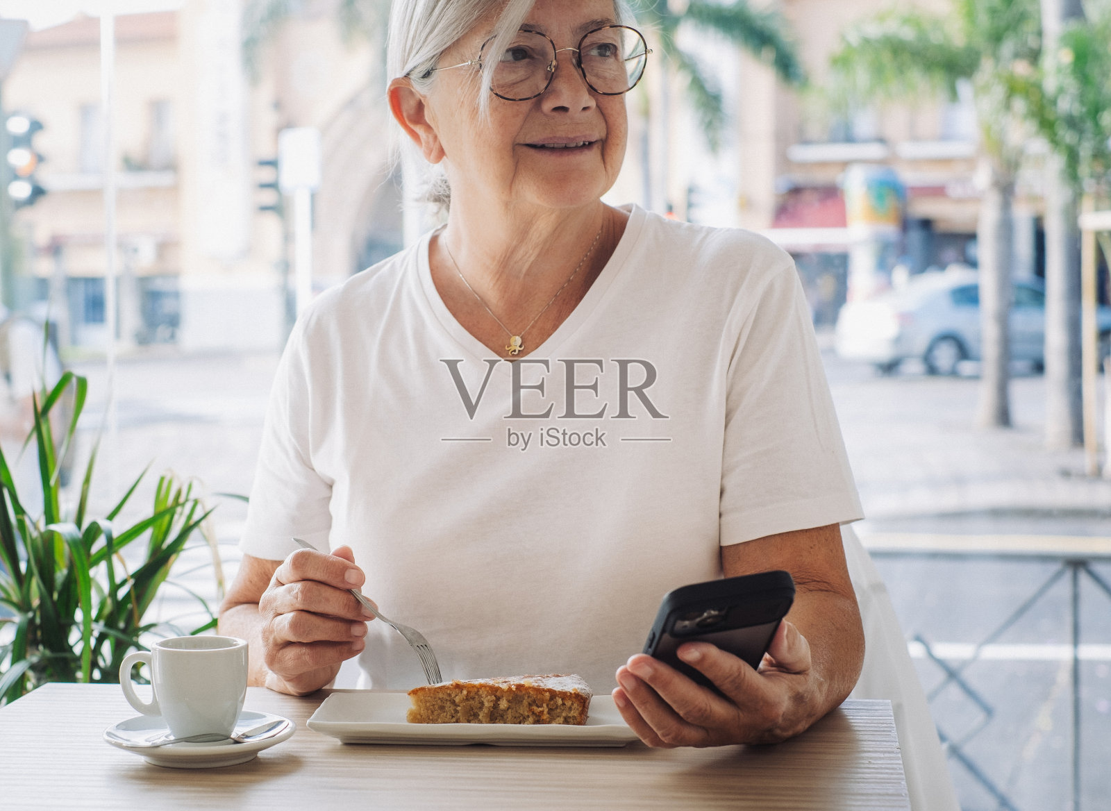 戴着眼镜、面带微笑的老年妇女坐在咖啡桌旁，手里拿着手机，用浓咖啡杯和一片杏仁蛋糕休息照片摄影图片