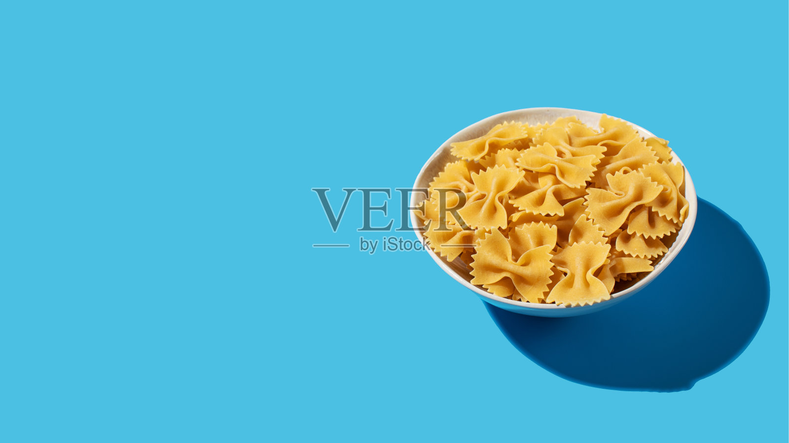 未煮熟的领结意面，在充满活力的蓝色背景下，落在白色碗里，完美的烹饪概念。本空间照片摄影图片