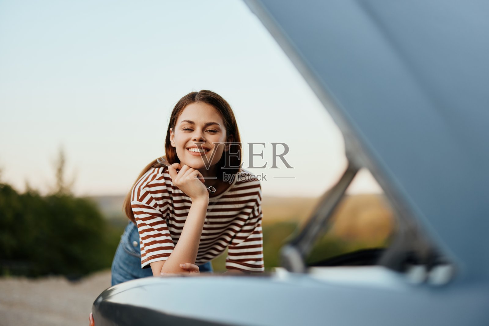 一个女人拿着扳手，微笑着开心地看着打开的引擎盖下，独自在公路旅行中修理路边故障的汽车照片摄影图片