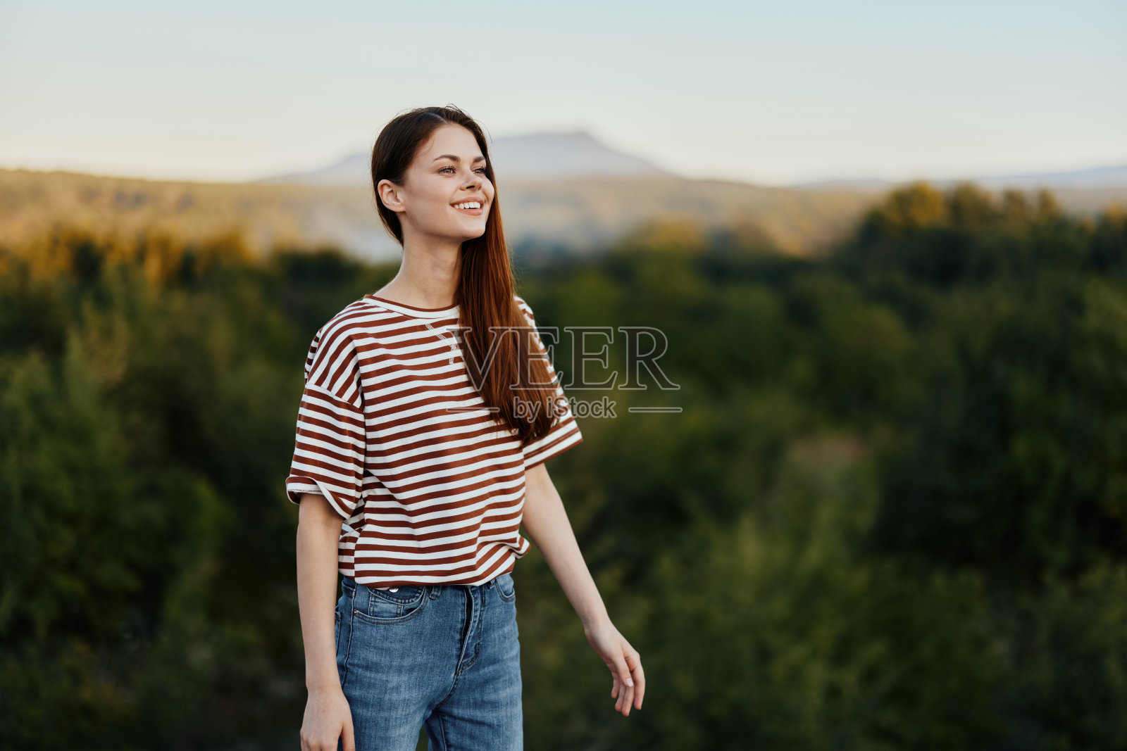 一个年轻女子笑着看着镜头，她穿着简单的衣服，背景是美丽的秋日山林。移动的生活方式照片摄影图片