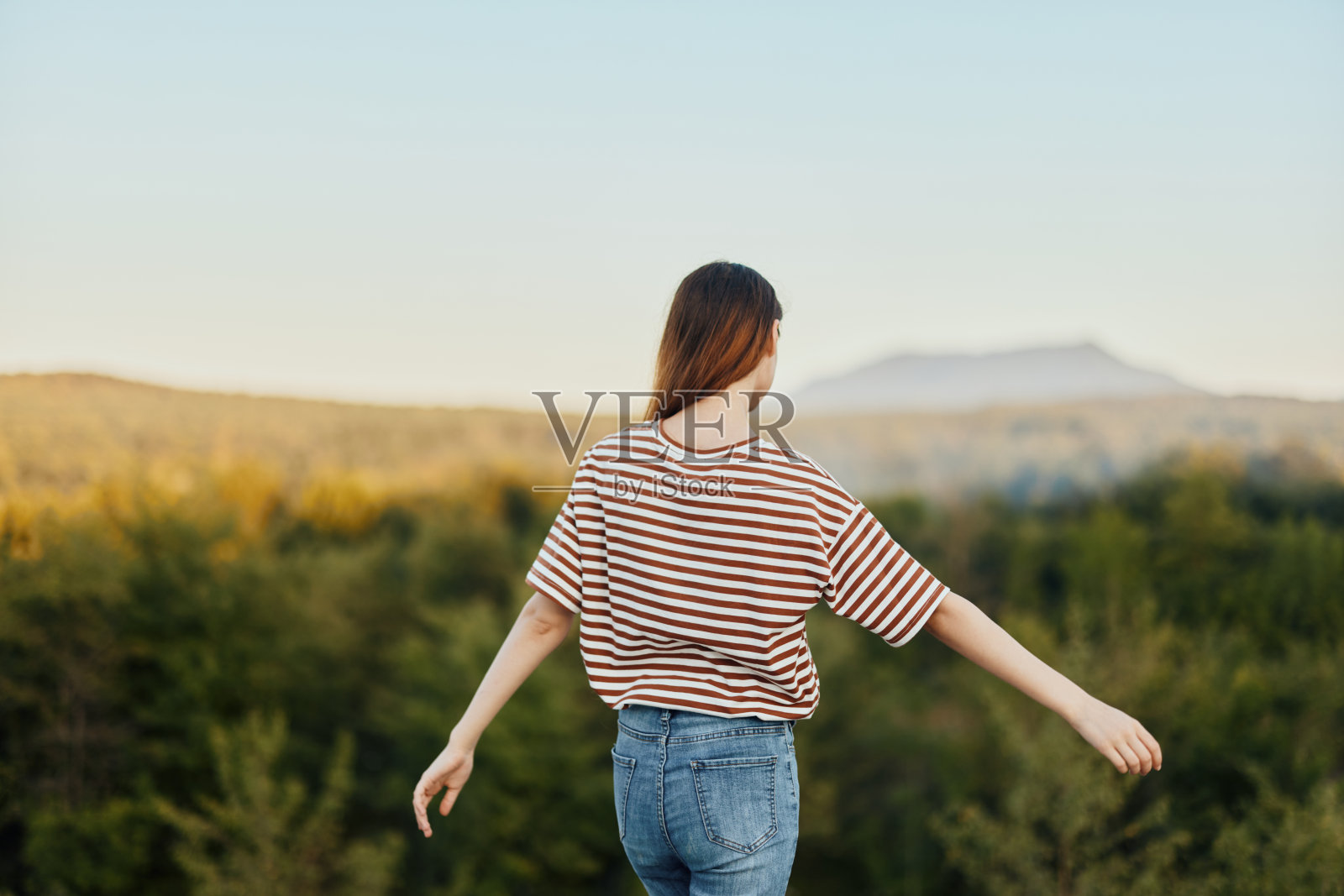 一名年轻女子身穿t恤和牛仔裤，背对着镜头站在大自然中，欣赏着美丽的山景。秋游自然生活方式照片摄影图片