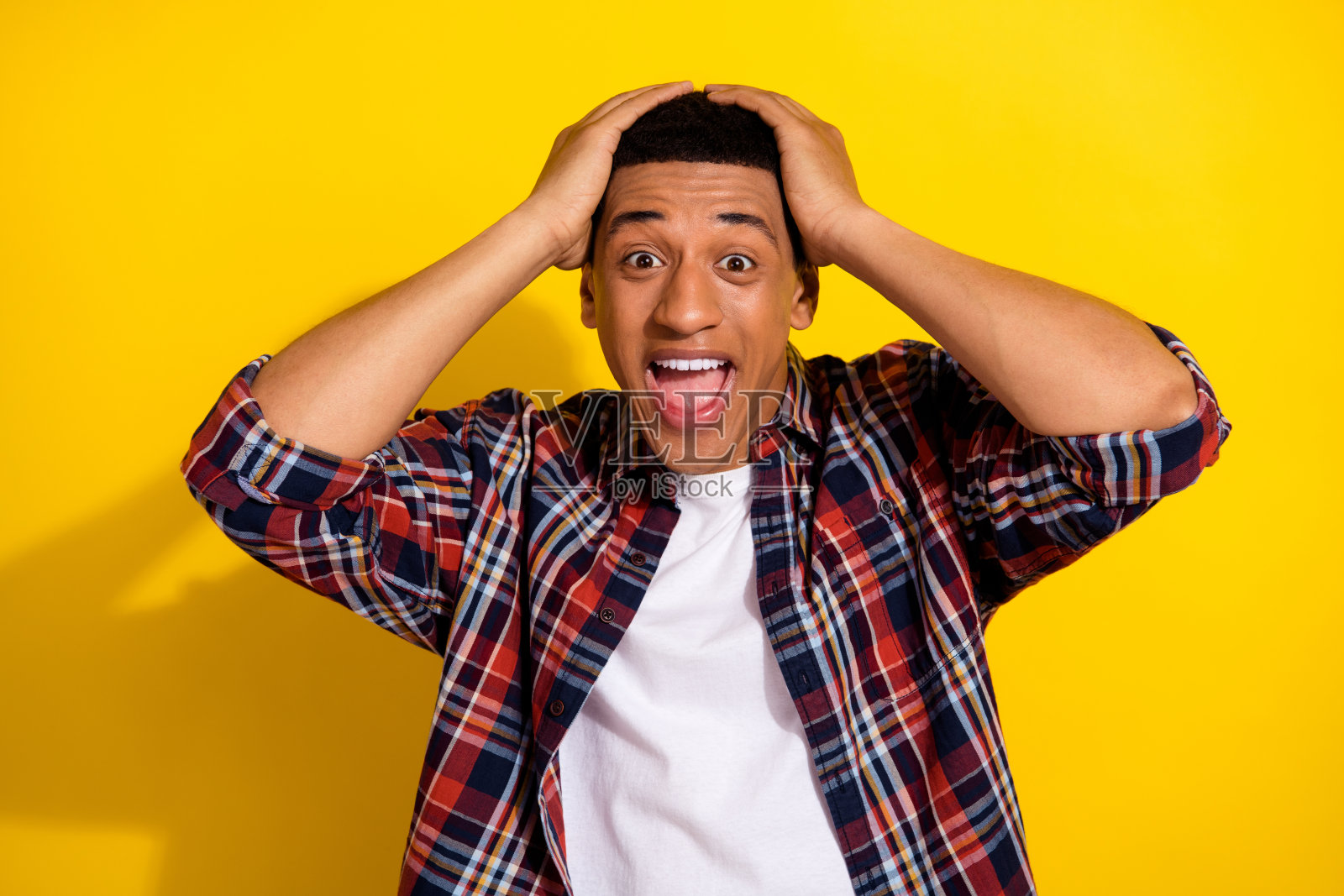照片中的男子穿着时髦的格纹衣服，手臂触摸头部，张开嘴巴，孤立在黄色背景上照片摄影图片