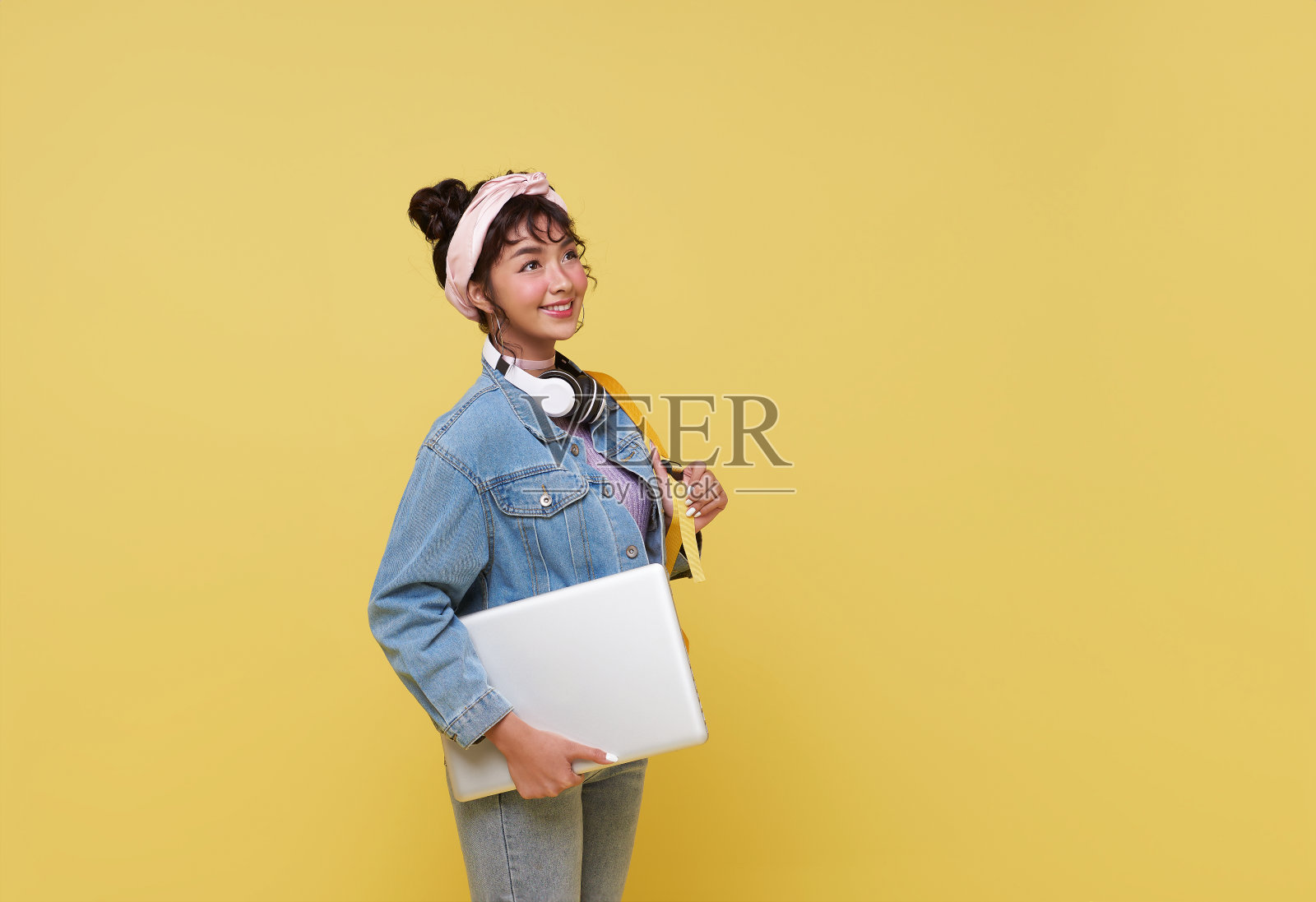 一个快乐微笑的年轻亚洲大学生的肖像，带着笔记本电脑和背包，准备在黄色背景上单独上学。照片摄影图片