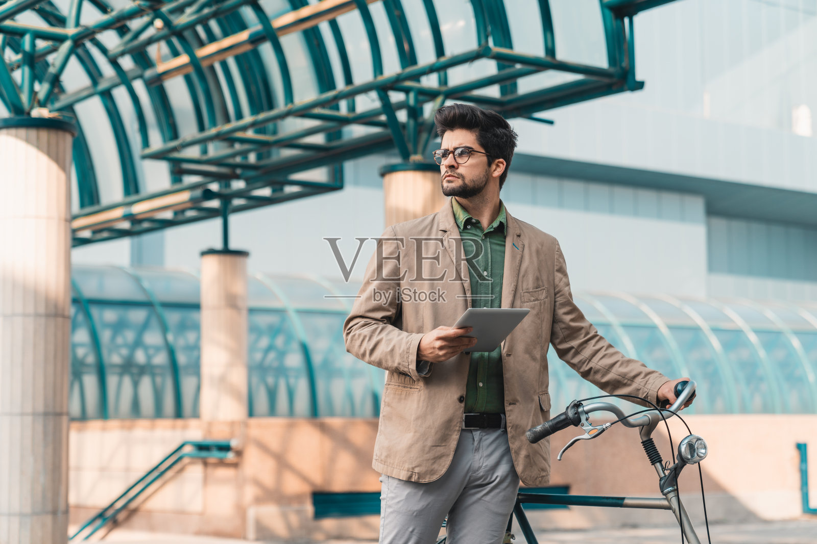 严肃的年轻企业家一边拿着平板电脑，一边站在自行车旁看向别处照片摄影图片