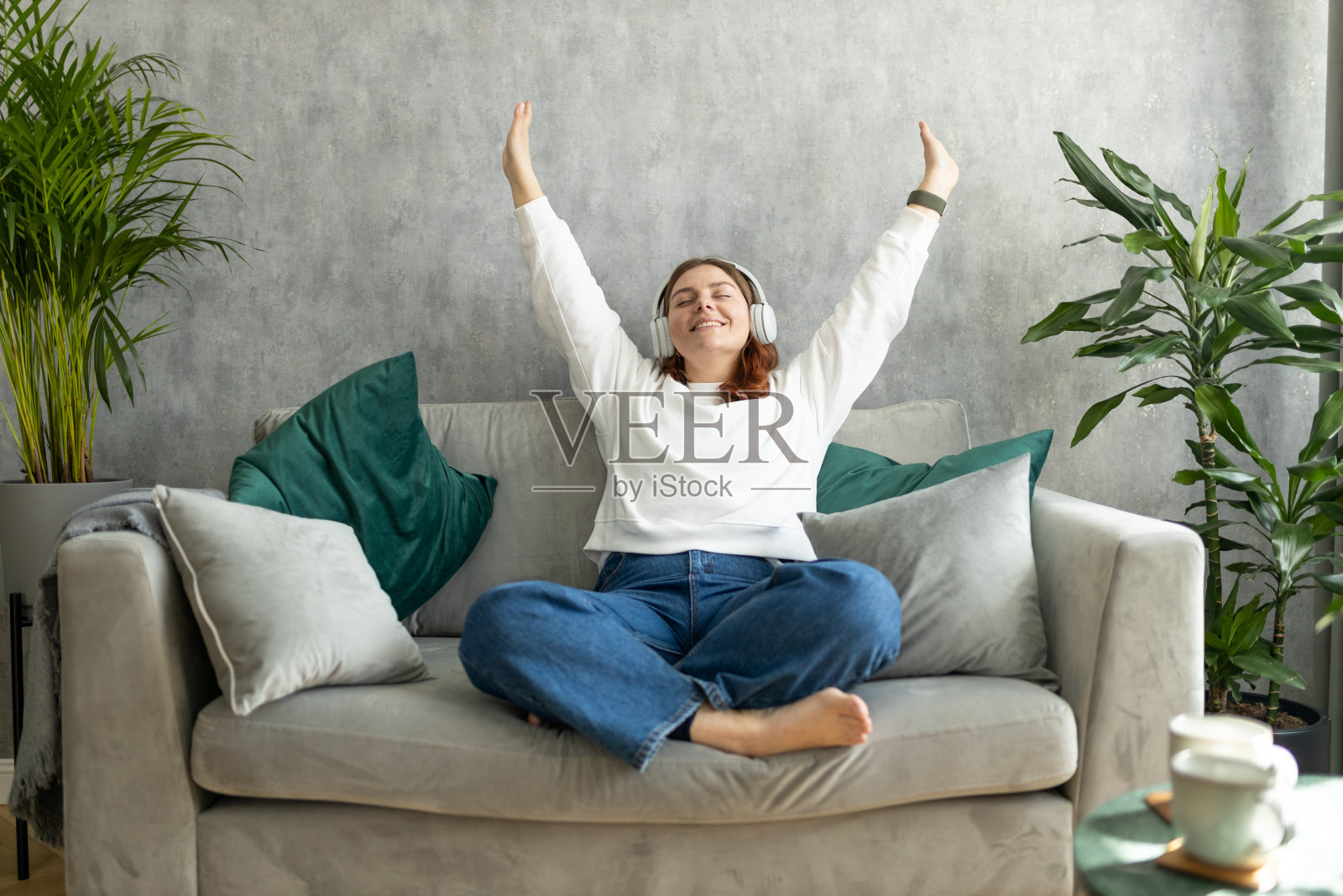 微笑的女孩在家里放松，她正在听音乐，用智能手机，戴着白色耳机。30多岁的年轻女性坐在家里的沙发上享受着良好的音响效果。全身照片摄影图片